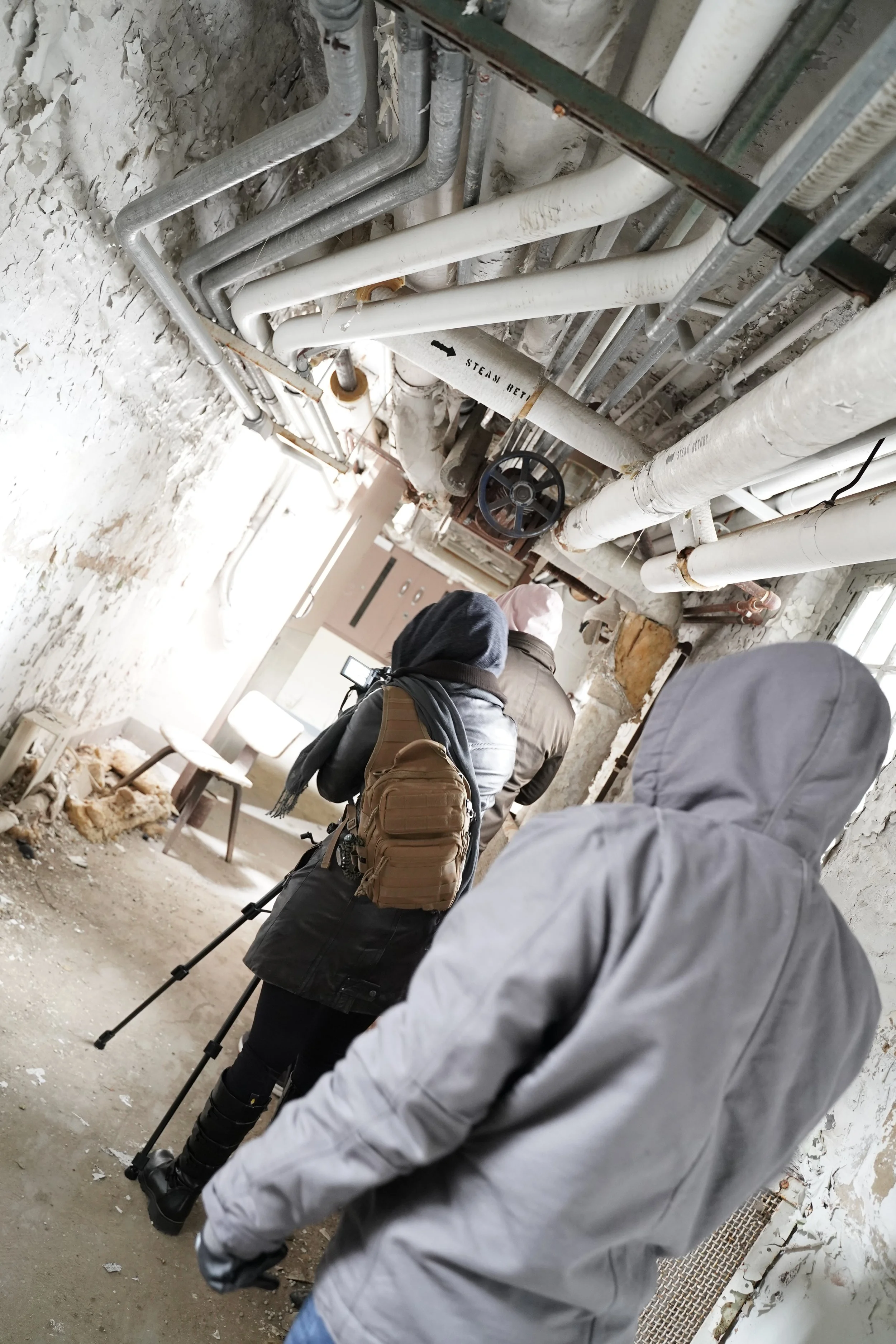 People in a construction or renovation site, with exposed pipes and a dirt floor, some carrying backpacks and using a camera or phone for taking pictures.