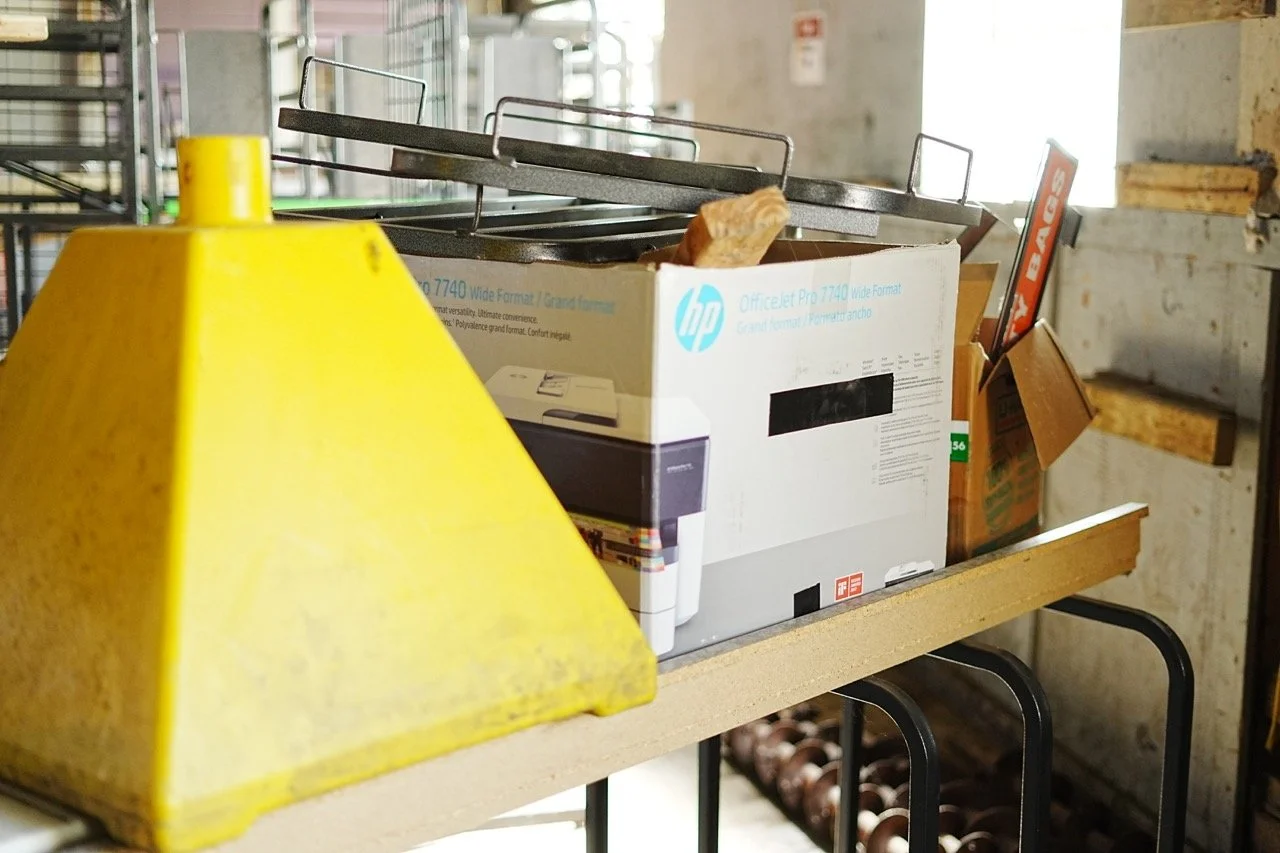 A yellow caution sign, stacked boxes including an HP printer box, and metal shelves with various items in an industrial or warehouse setting.