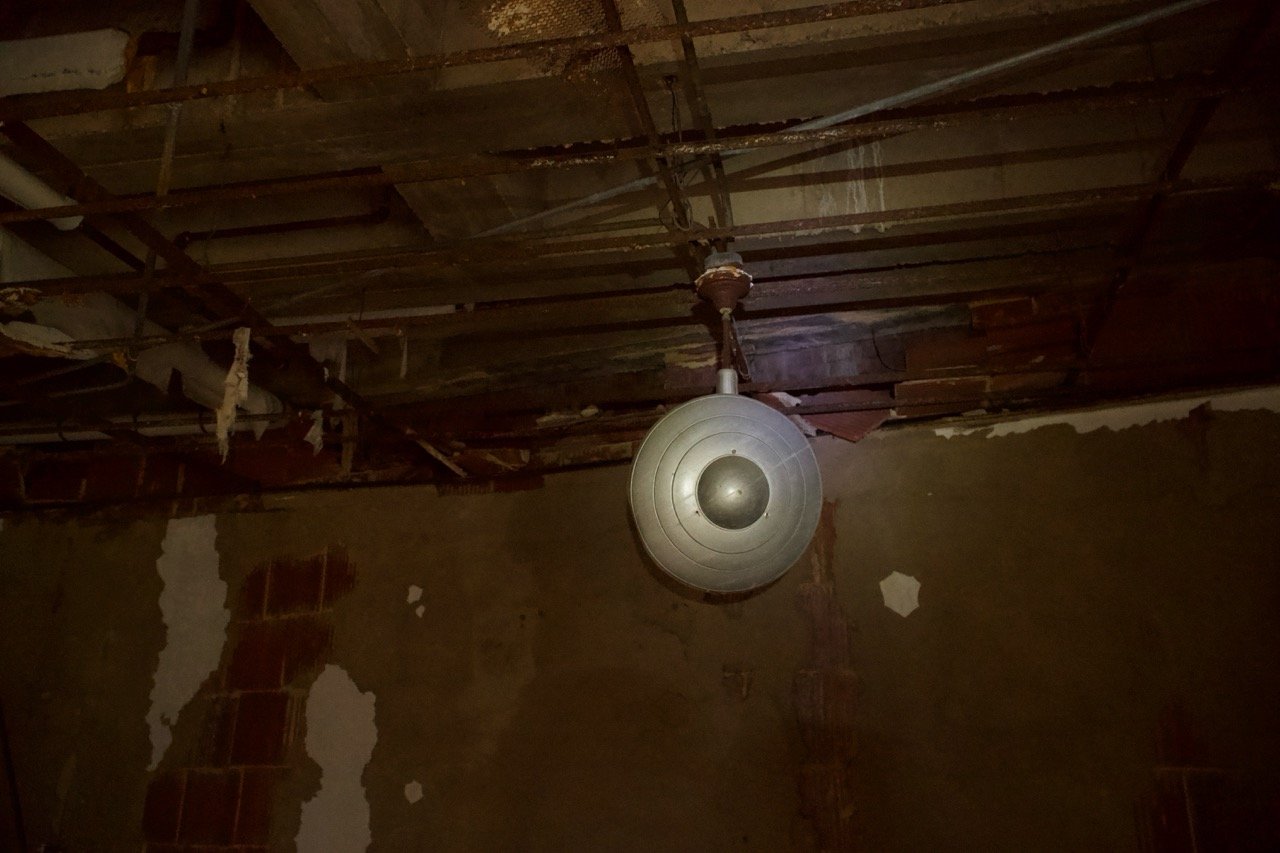 A vintage ceiling light fixture hanging from a damaged ceiling with exposed wood and pipes in a dilapidated room.