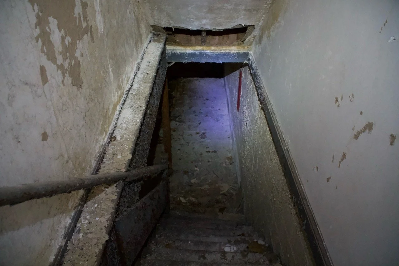 A dark, narrow stairwell under construction or renovation with exposed, worn steps, peeling paint on the walls, and a visible hole in the ceiling at the top.