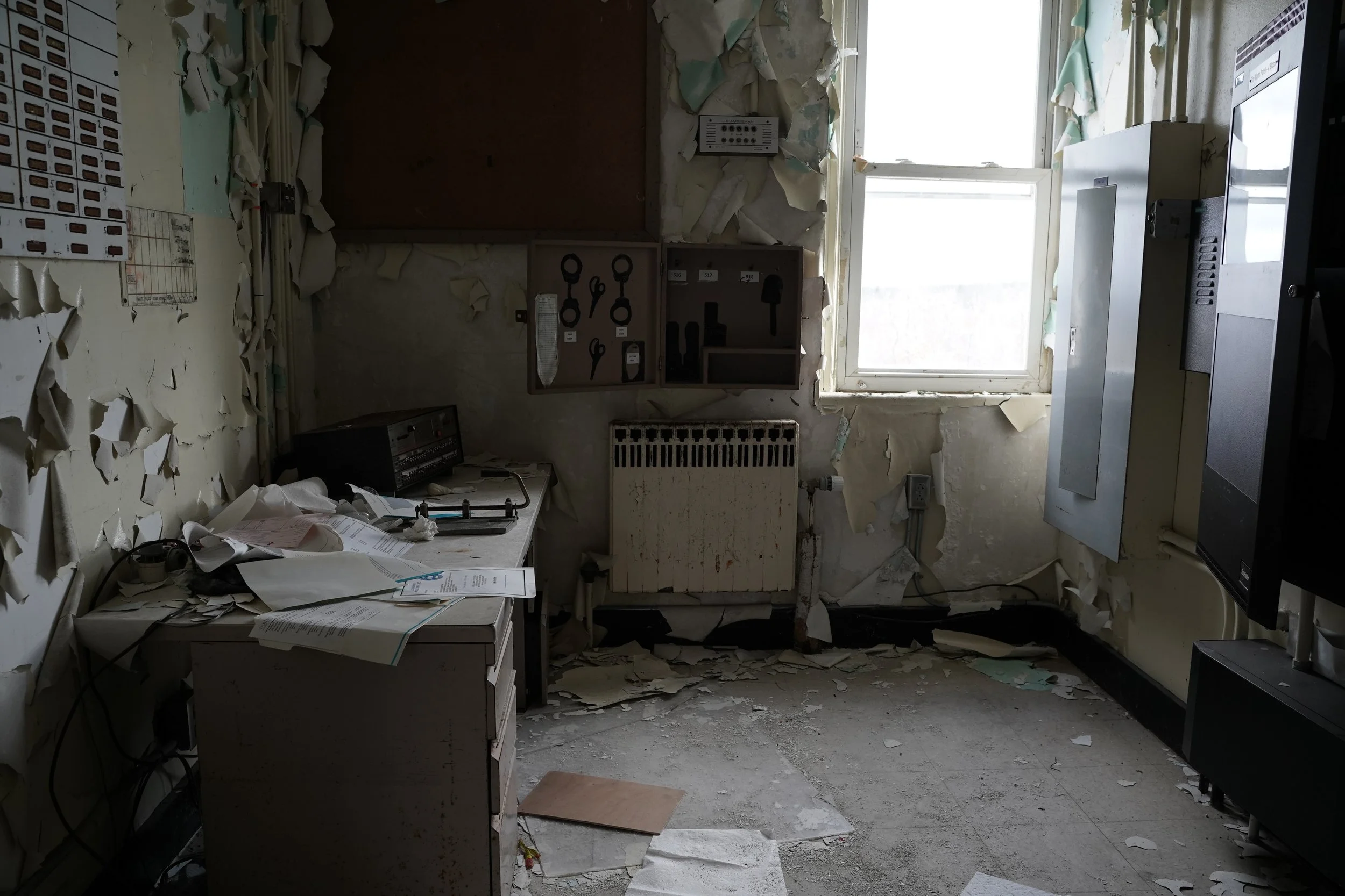 A cluttered and dilapidated office or utility room with peeling paint on the walls, scattered papers and wires, a window with broken or missing panes, and various equipment and tools. The room appears abandoned and in disrepair.