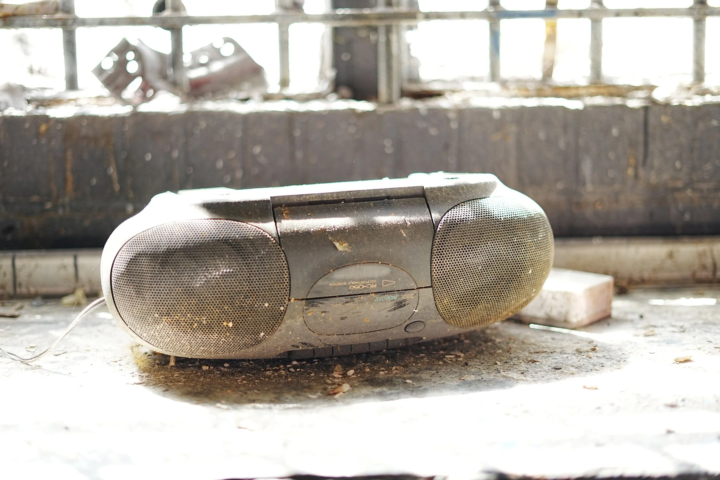 An old, dusty portable stereo lying on a dirty concrete surface with debris and dirt around, in a rustic, possibly abandoned, setting with a weathered background.