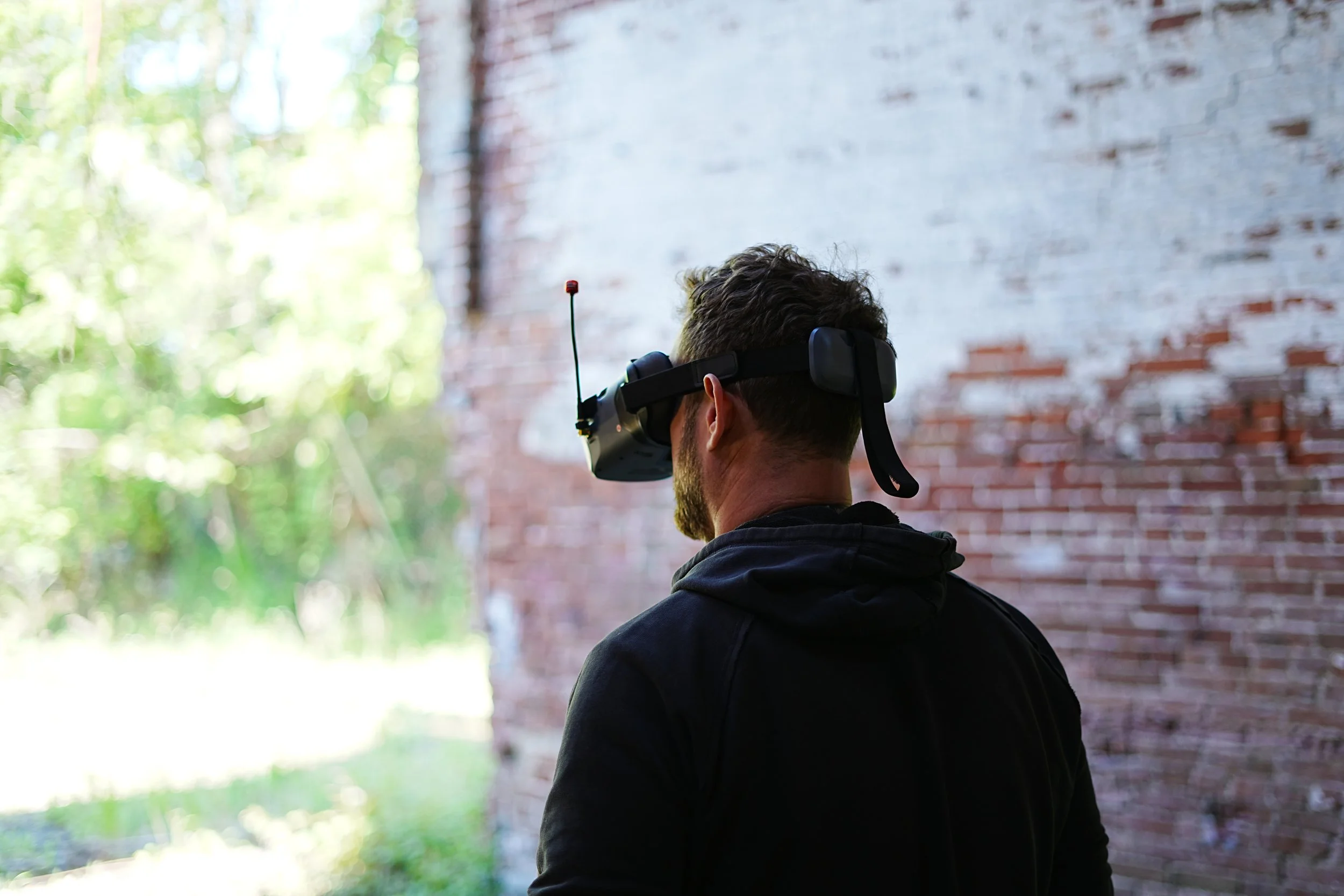 A person wearing a virtual reality headset, standing indoors near a brick wall with outdoor scenery visible through windows.