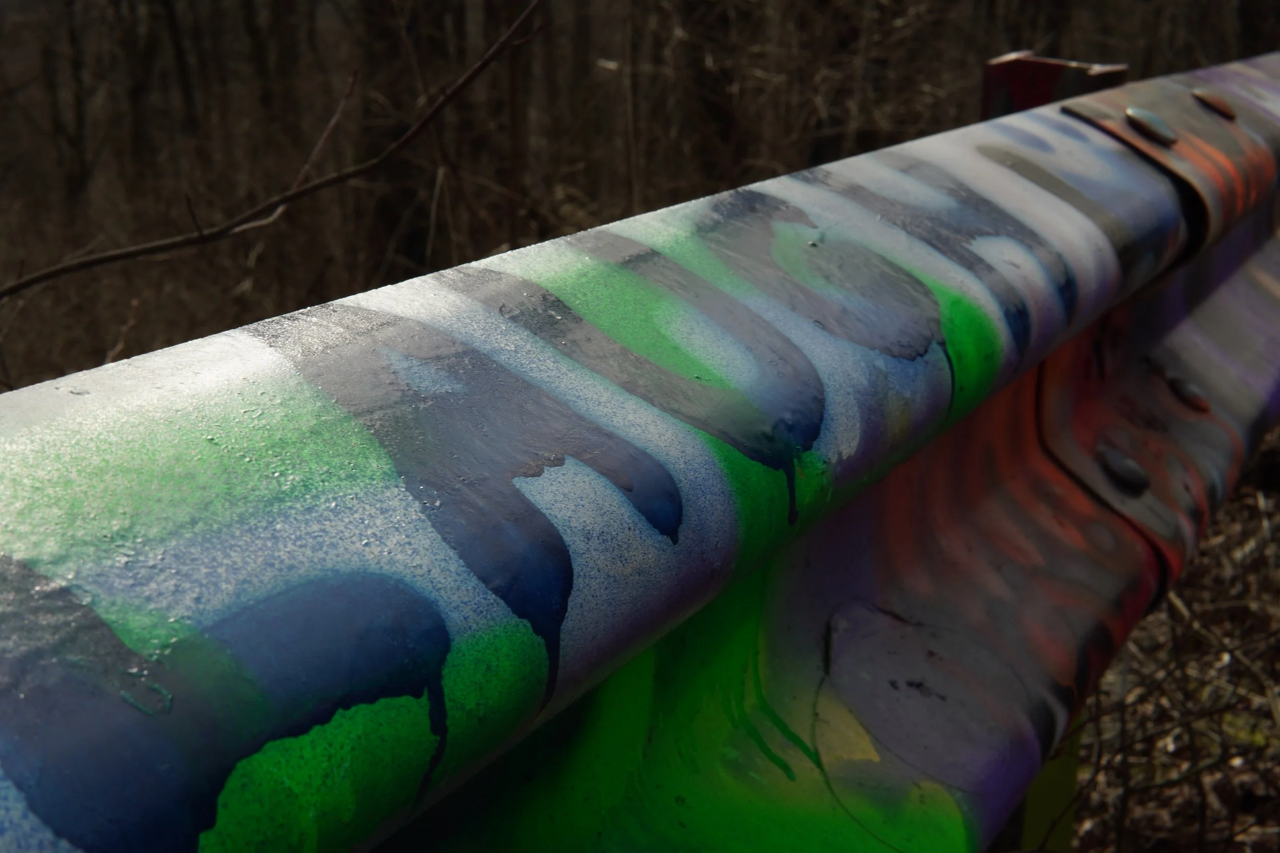 Close-up of a graffiti-covered metal pipe with green, blue, and purple paint, situated outdoors among dry grass and twigs.