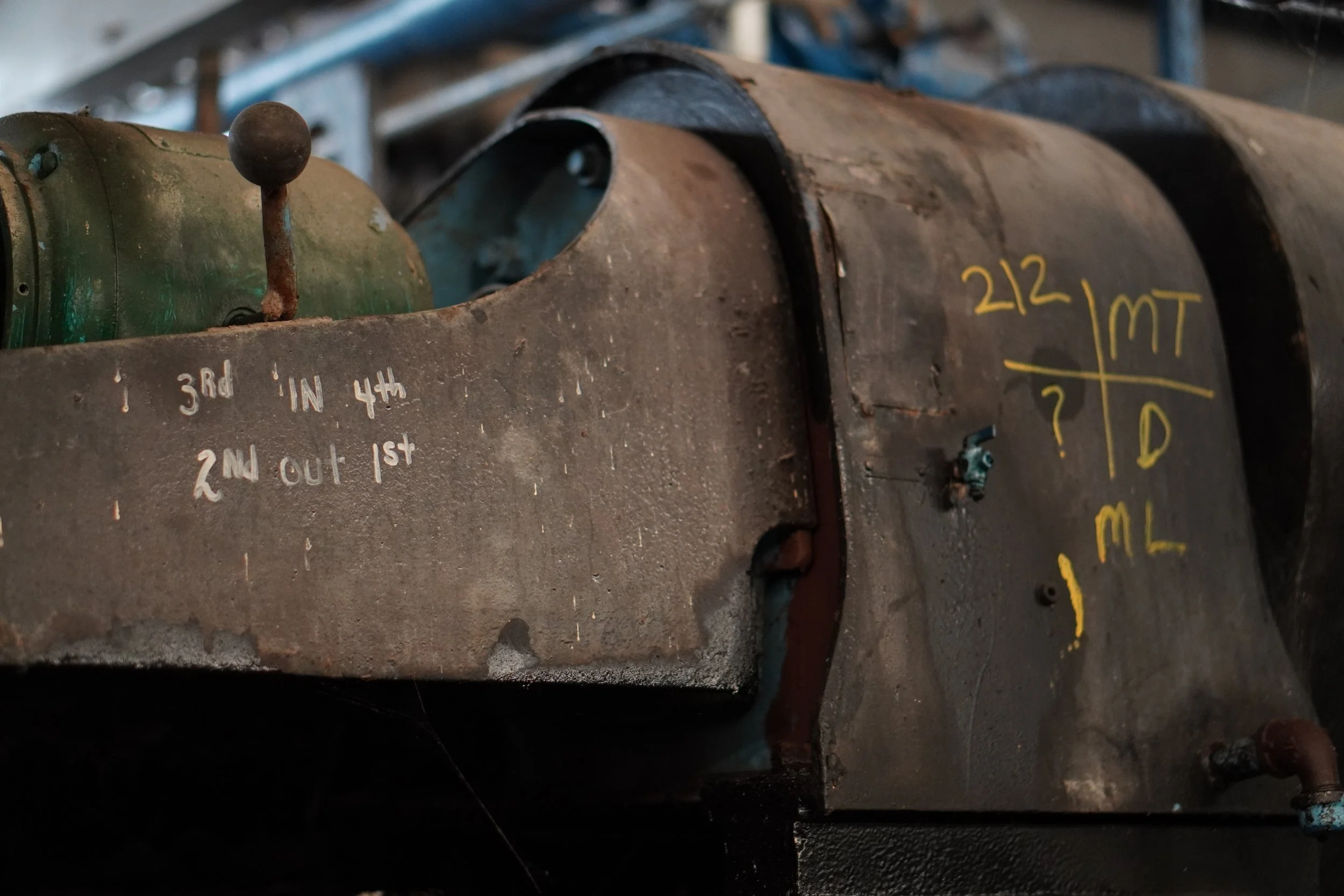 Close-up of a rusty, cylindrical mechanical component with handwritten markings in yellow and white, including the numbers '212,' and instructions such as '3rd in, 4th out, 1st.' The background is blurred with a metallic structure.