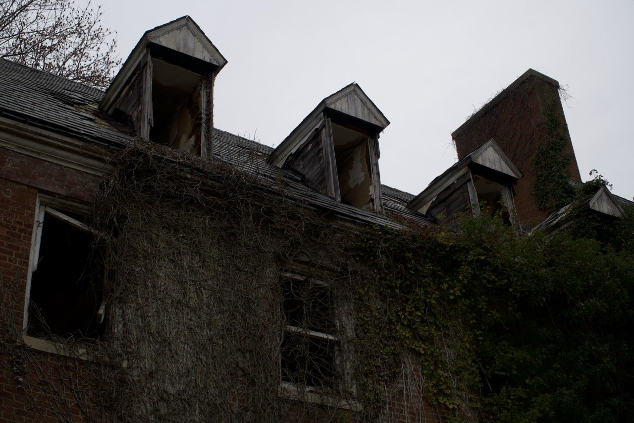 An old, abandoned brick building with multiple broken windows and boarded-up attic dormer windows. The roof is damaged and overgrown with vines and weeds.