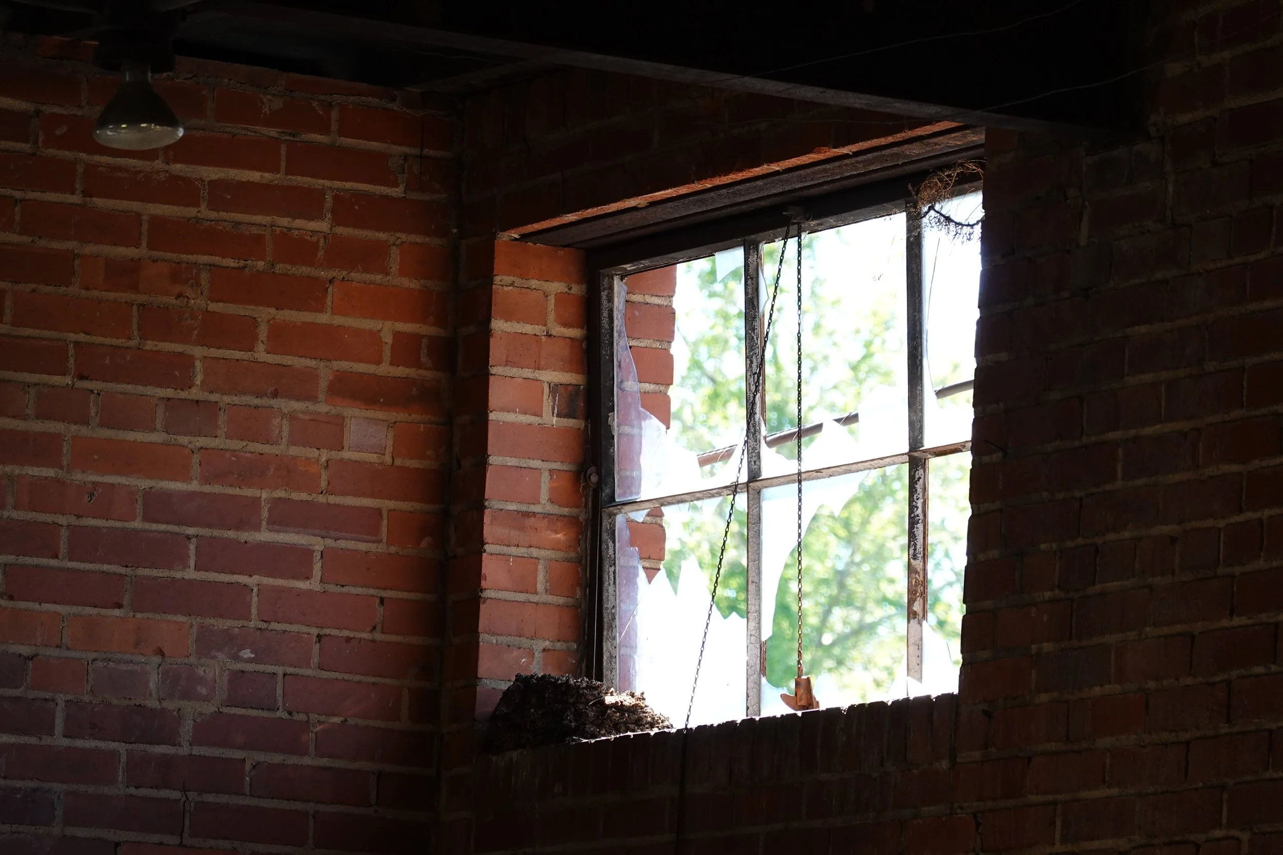 An unfinished brick wall with a large window opening, with chains hanging inside, and sunlight filtering through the window to the outside.