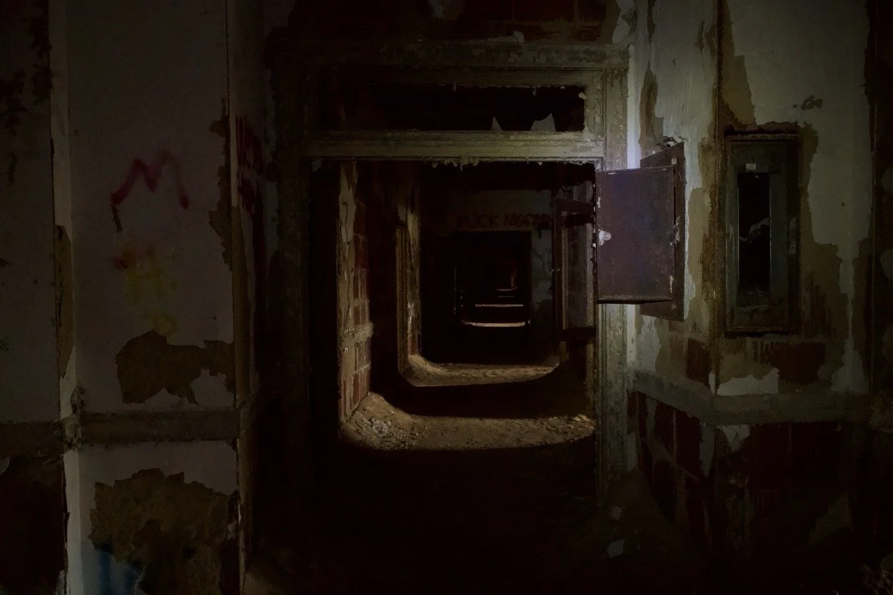 Dark, abandoned hallway with peeling walls and open electrical boxes.