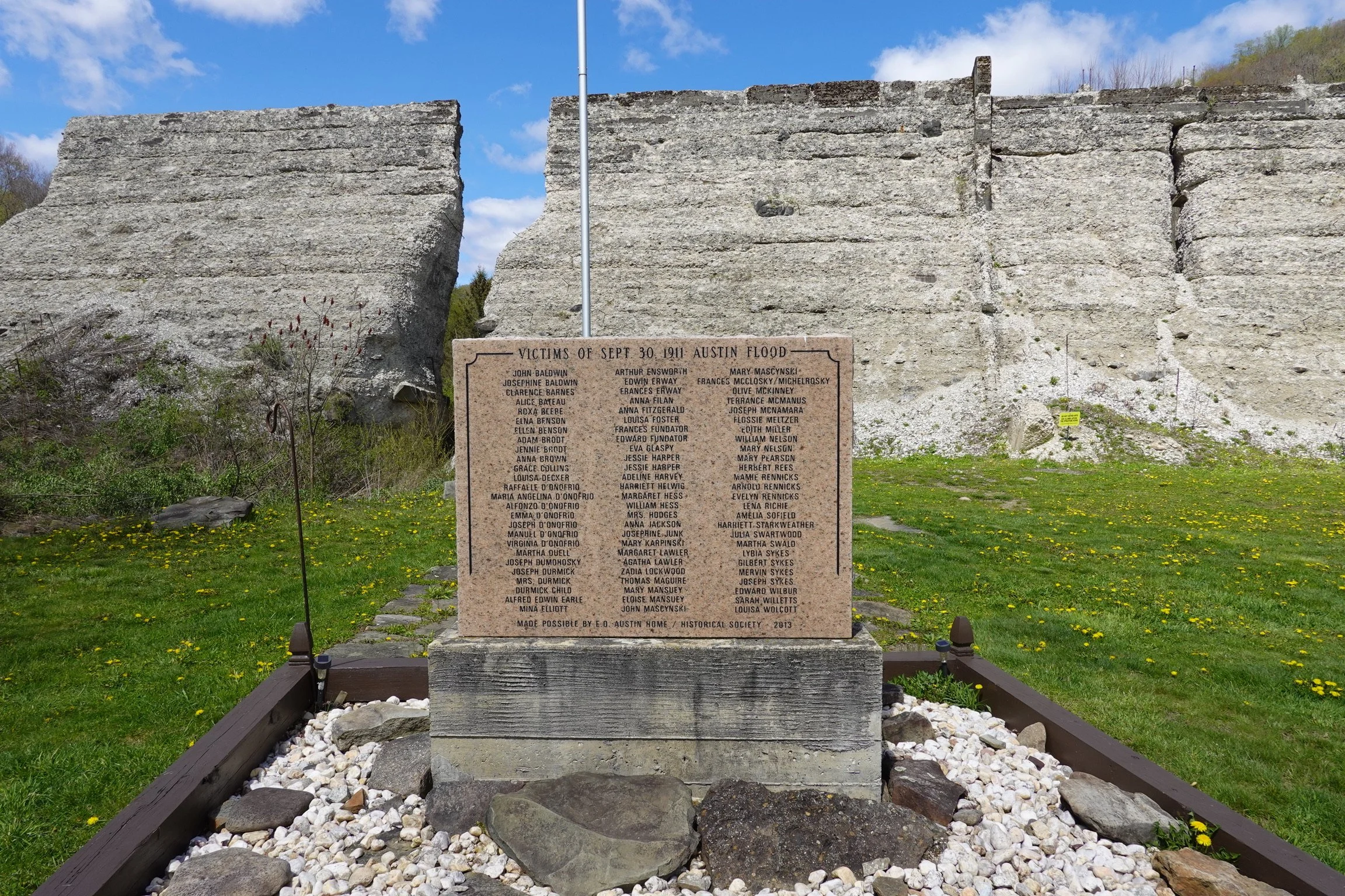 A memorial stone honoring victims of the September 30, 1911 Austin flood, with a list of names and a caption at the bottom. The stone is set on a grassy area with yellow flowers, surrounded by a wooden frame, and the sky is partly cloudy with trees i