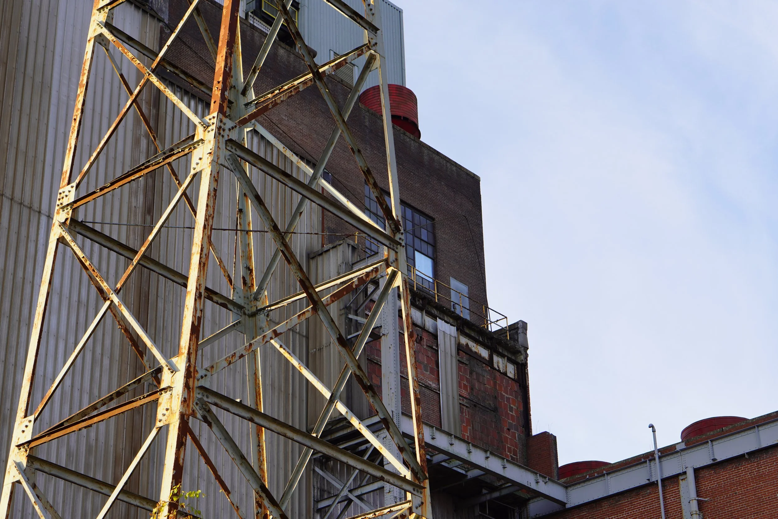 An old rusty metal tower in front of an industrial brick building with metal siding. The sky is clear and blue.