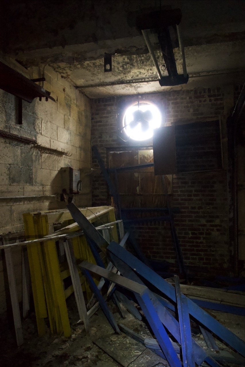 A dimly lit, cluttered basement or garage with exposed brick and concrete walls, scattered blue and yellow metal shelving units, and a bright circular light fixture on the ceiling illuminating the space.