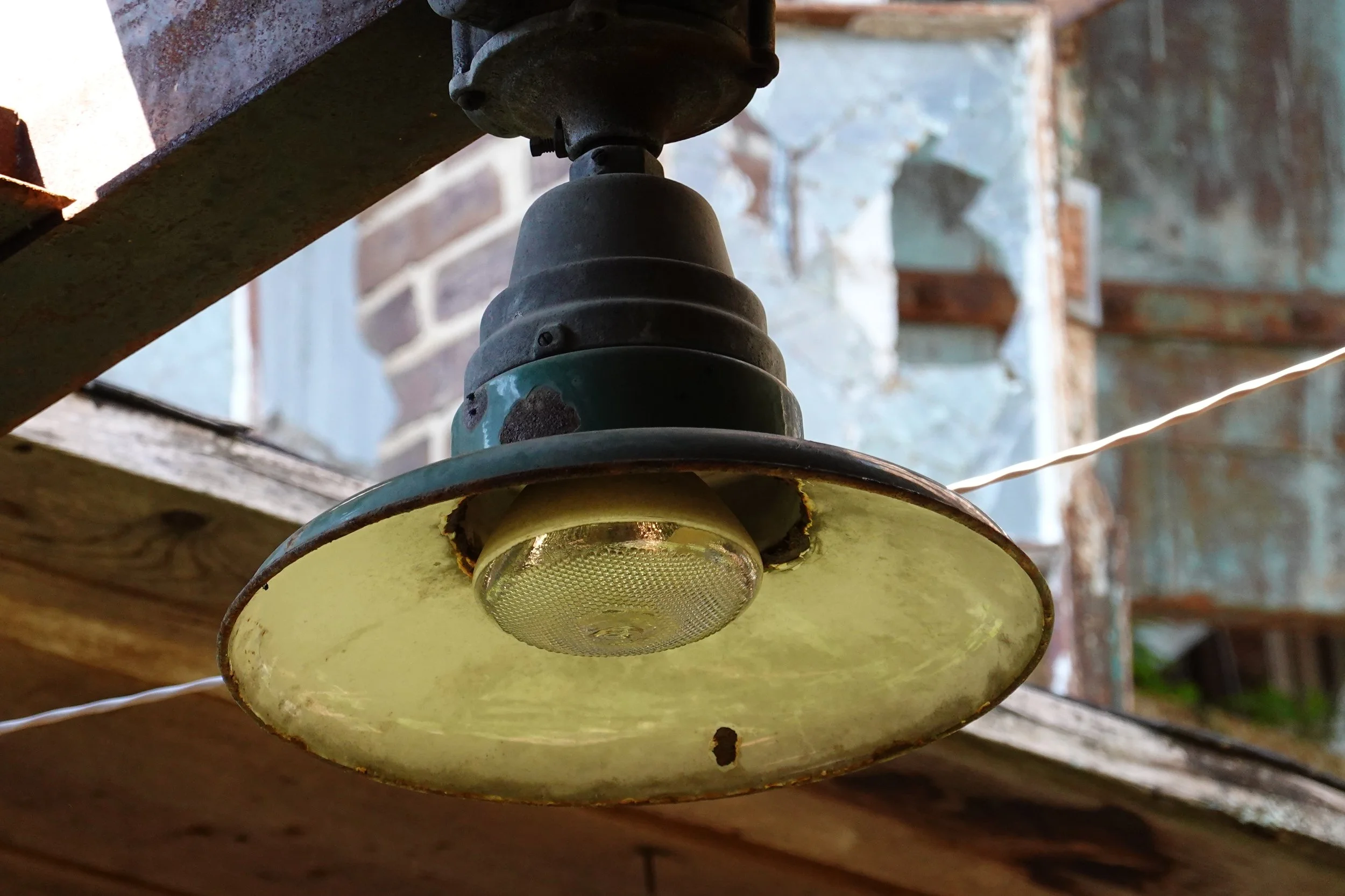 Close-up of an old, rusty outdoor hanging light fixture with a yellow glass shade, mounted under a wooden structure against a background of a brick wall and greenery.