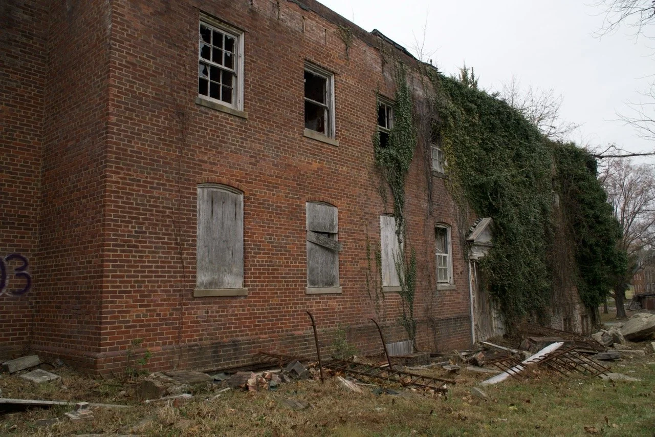 An abandoned brick building with boarded-up windows, some broken windows, and overgrown ivy. Debris and fallen metal objects are scattered in the front yard.