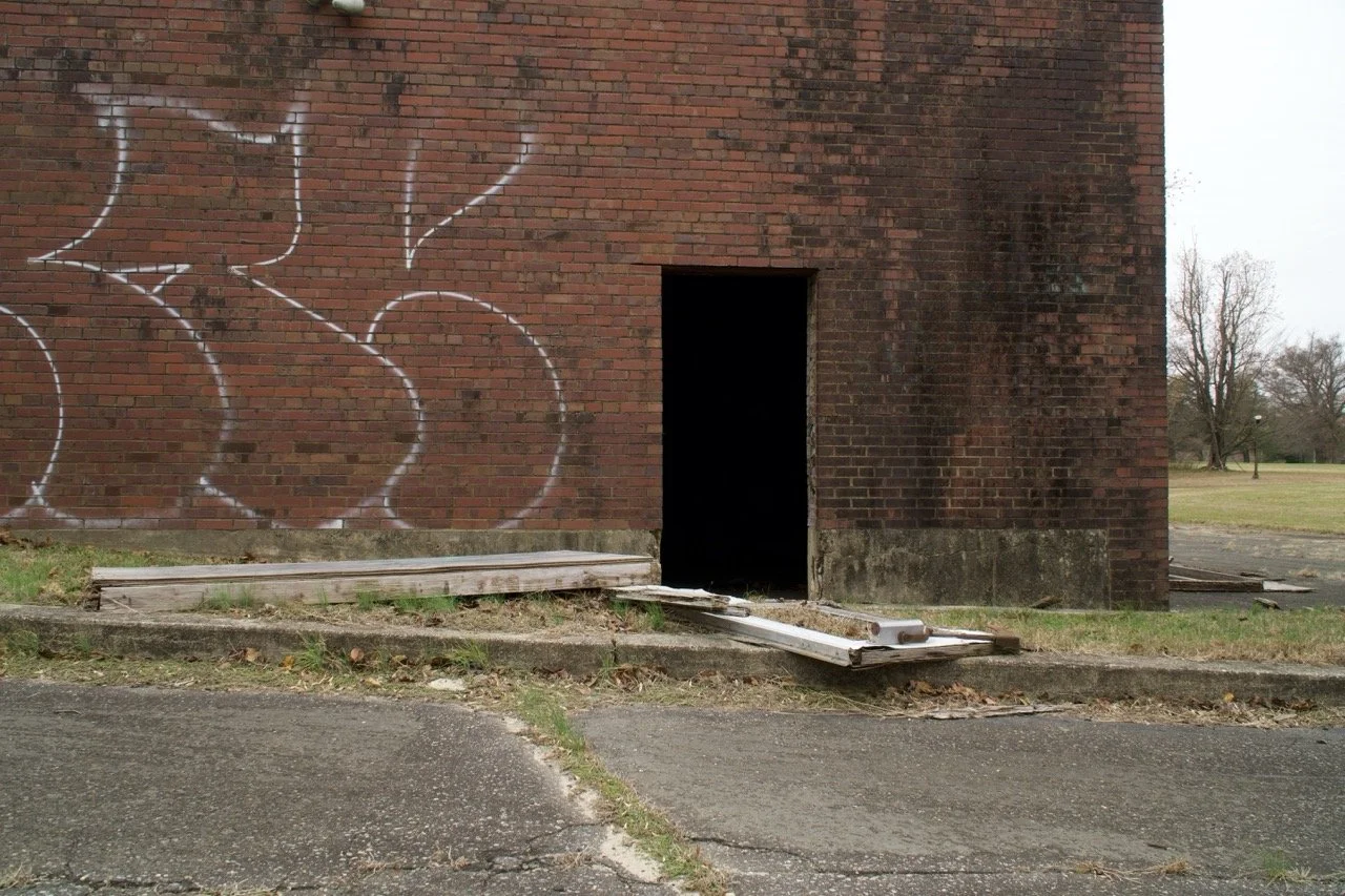 A brick building with graffiti on the wall, an open doorway, and a wooden ramp leading into it. The building is in a park-like area with grass and trees in the background.