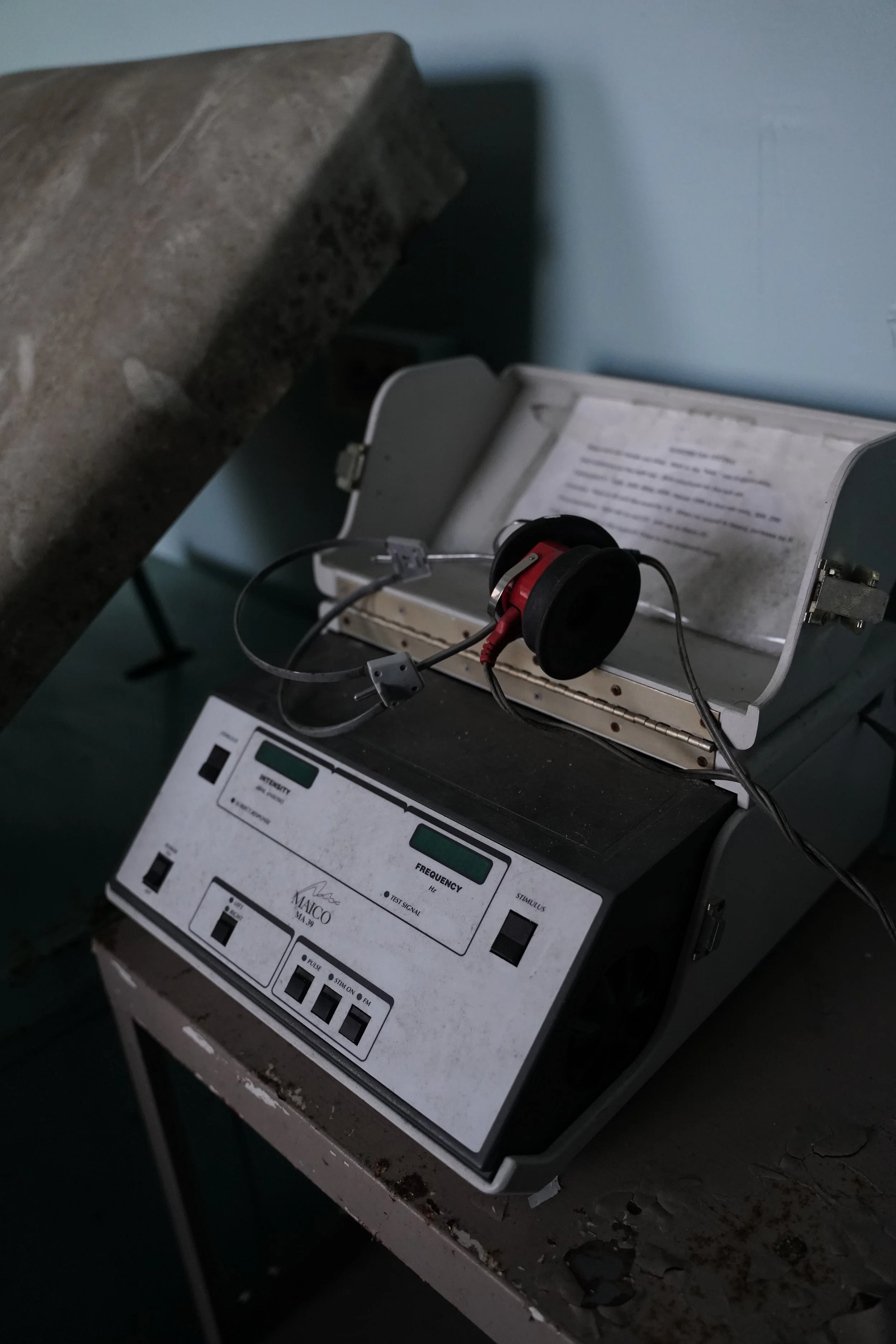 An old laboratory device with wires and a control panel, placed on a worn table with peeling paint. The device appears to be a scientific instrument with a metal frame, connected to a red and black headset, against a blue wall.