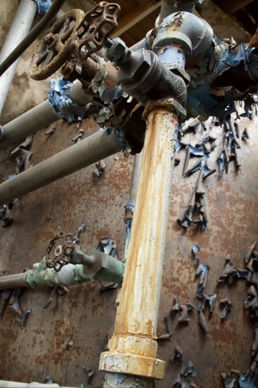 Corroded pipes with rusty valves and peeling paint in a deteriorated plumbing setup.