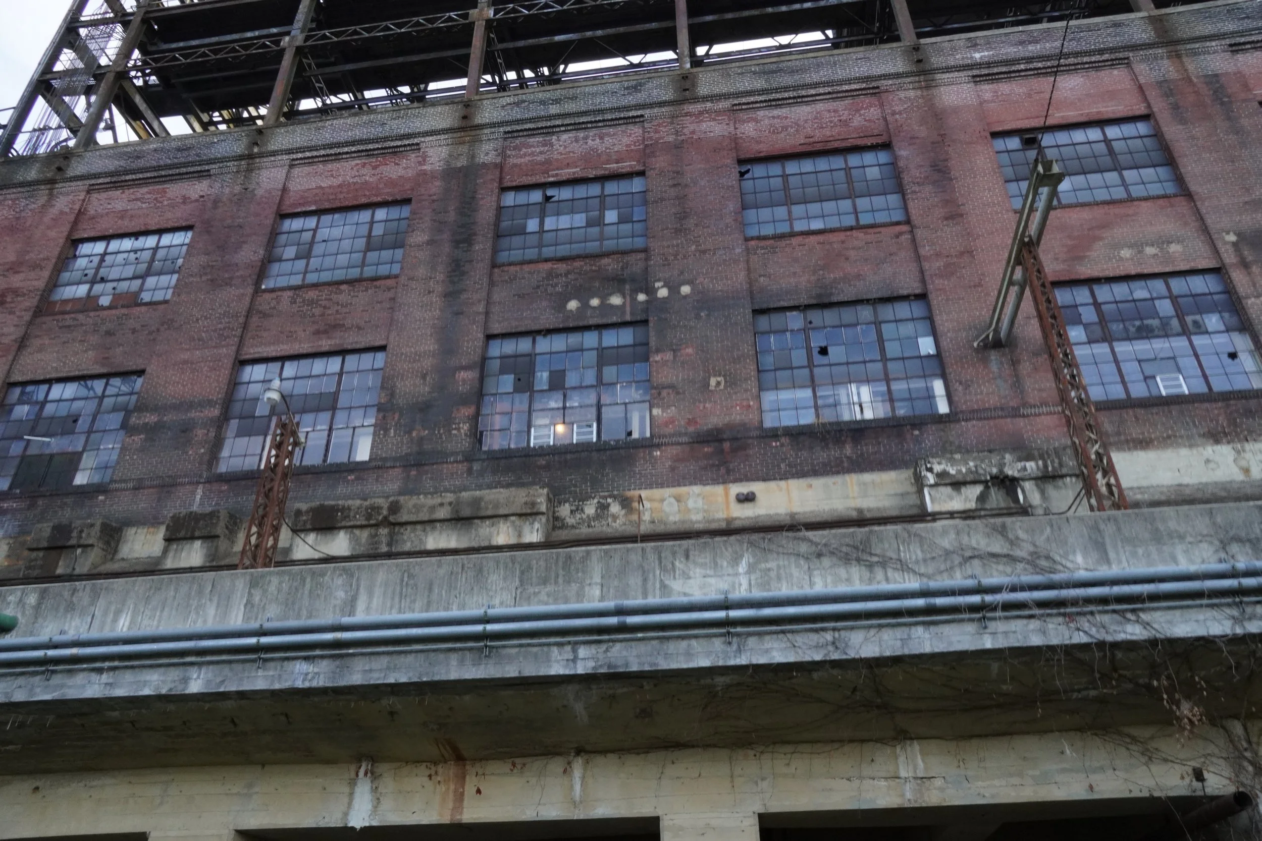 Close-up of an old brick industrial building seen from underneath a bridge with metal pipes in the foreground.