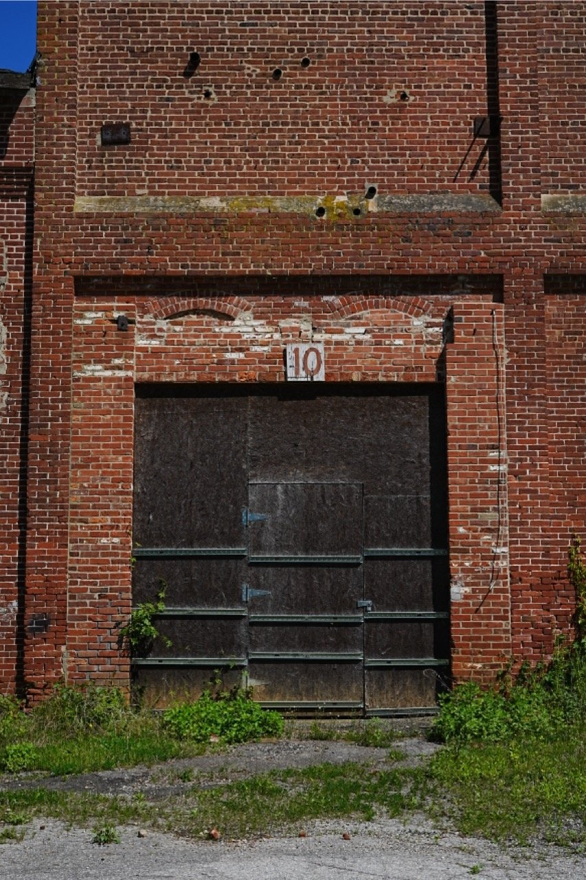 A large, old brick building with boarded-up windows and a metal garage door, with peeling bricks and graffiti, number 10 above the door, and overgrown plants at the base.