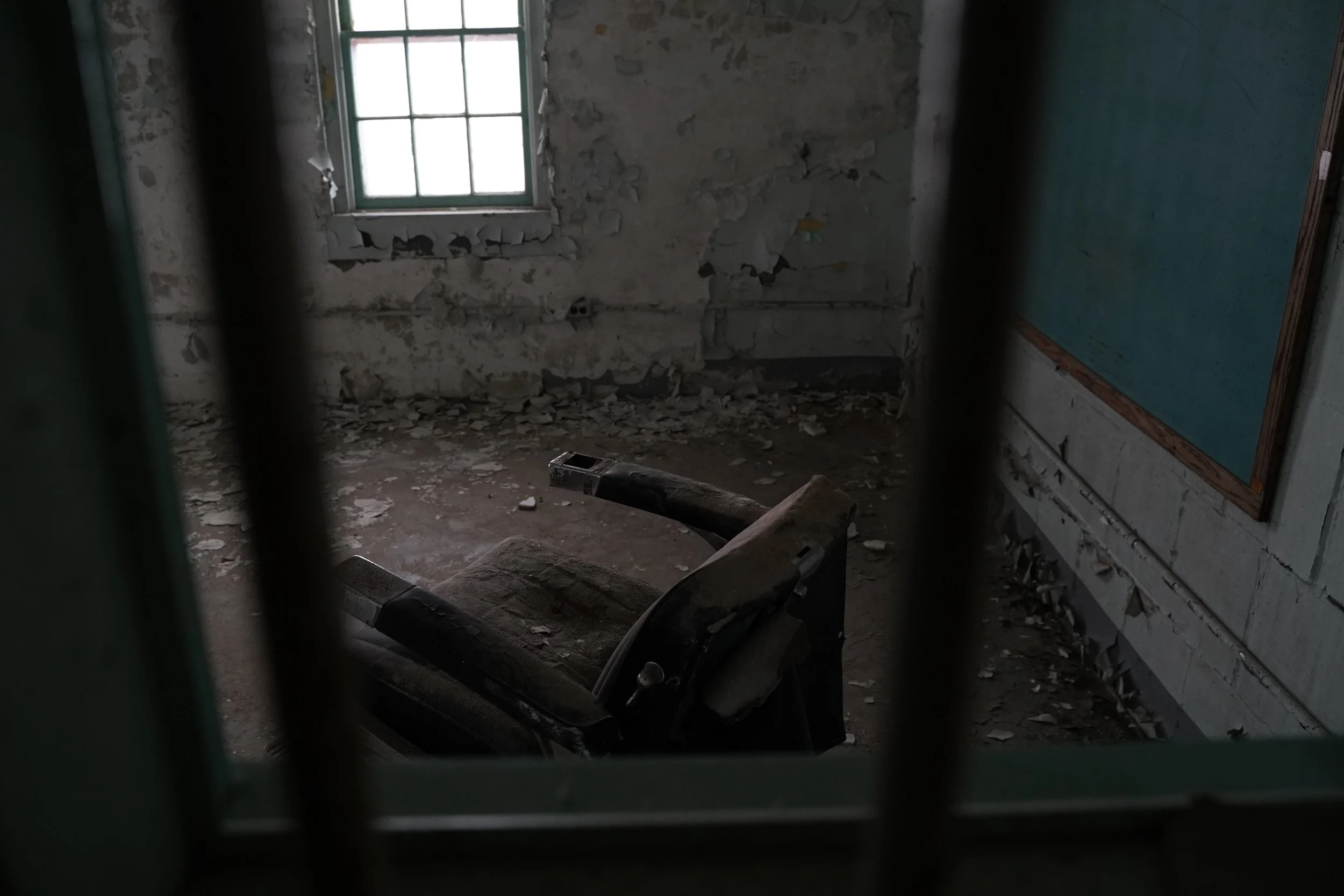 Room in a state of disrepair with peeling wall paint, debris on the floor, and an old, broken chair. A window with bars is visible in the lit background.