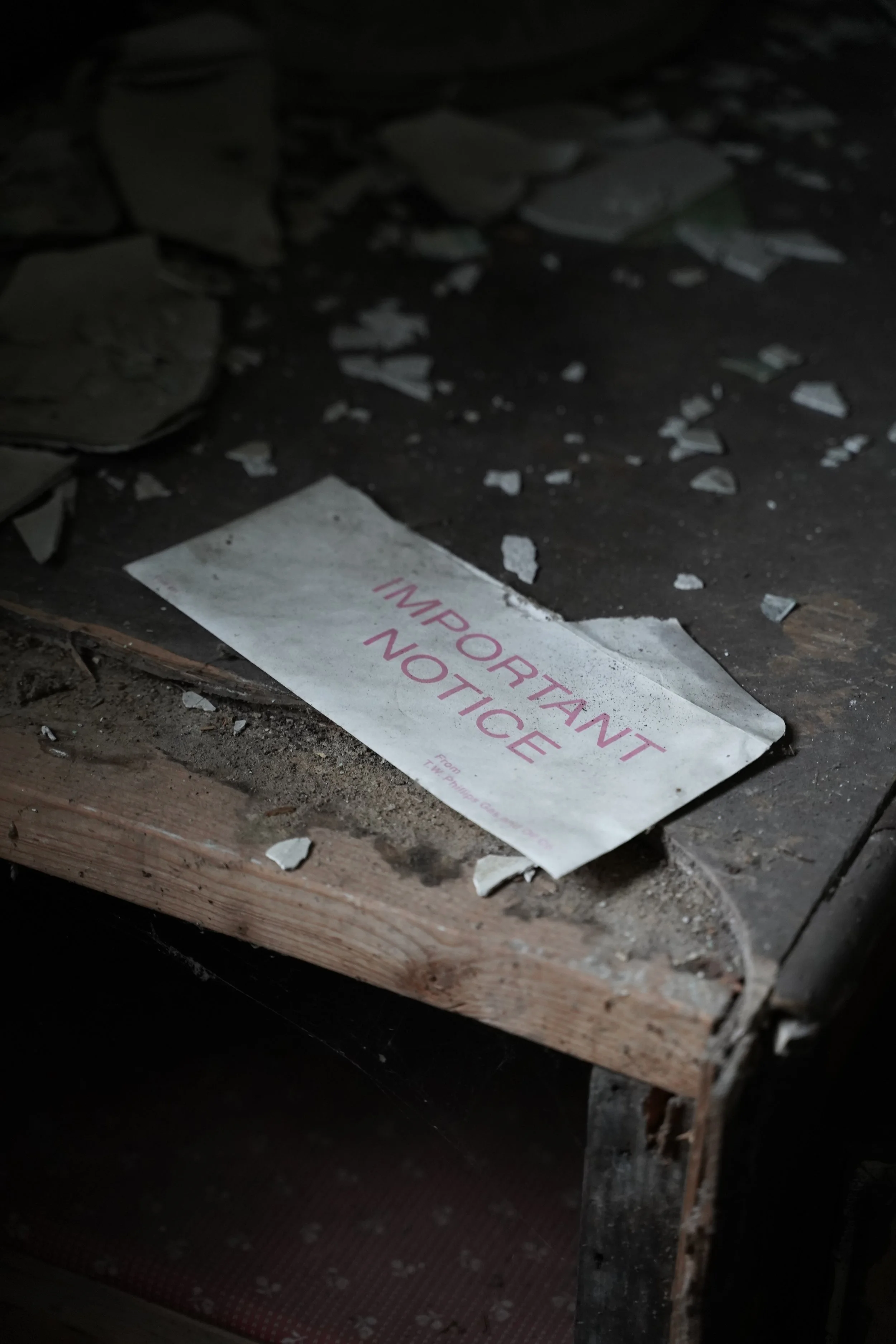 A broken sign on a dusty wooden surface that reads 'IMPORTANT NOTICE' surrounded by debris and broken plaster, with part of a red patterned floor visible below.