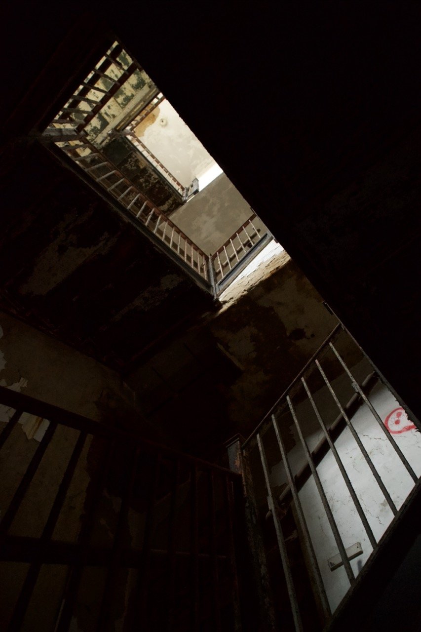 View of an abandoned, dark stairwell with peeling walls and metal railings, looking up towards a light source at the top.