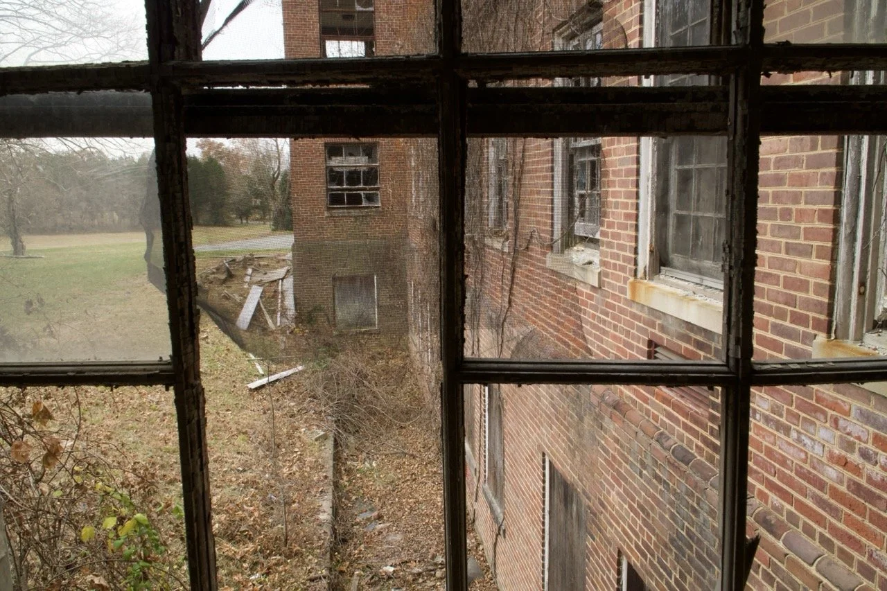 View through a dirty, broken window showing an abandoned brick building with damaged and boarded-up windows, and debris on the ground outside.