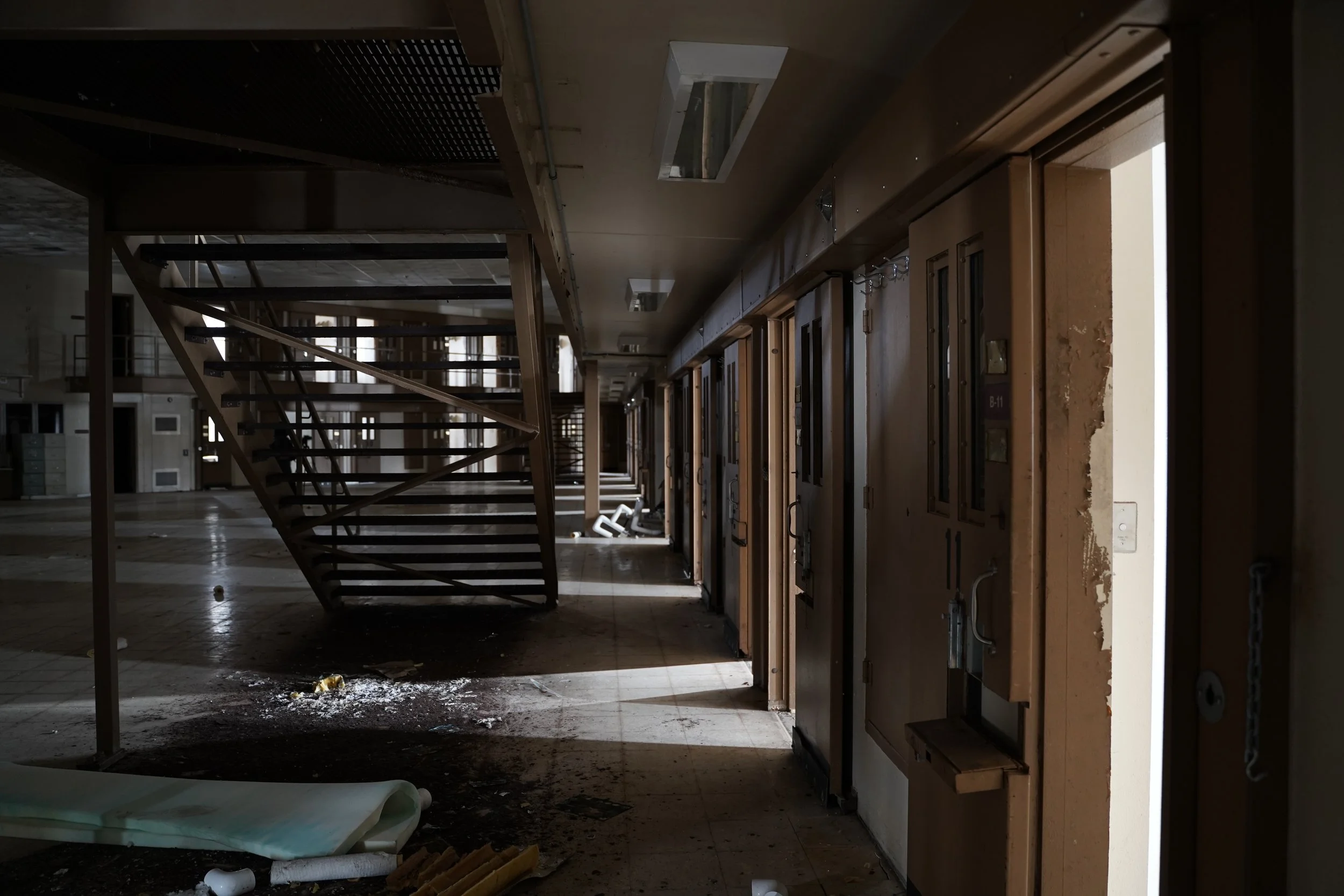 An abandoned hospital hallway with a staircase, empty room doors, and debris scattered on the floor.