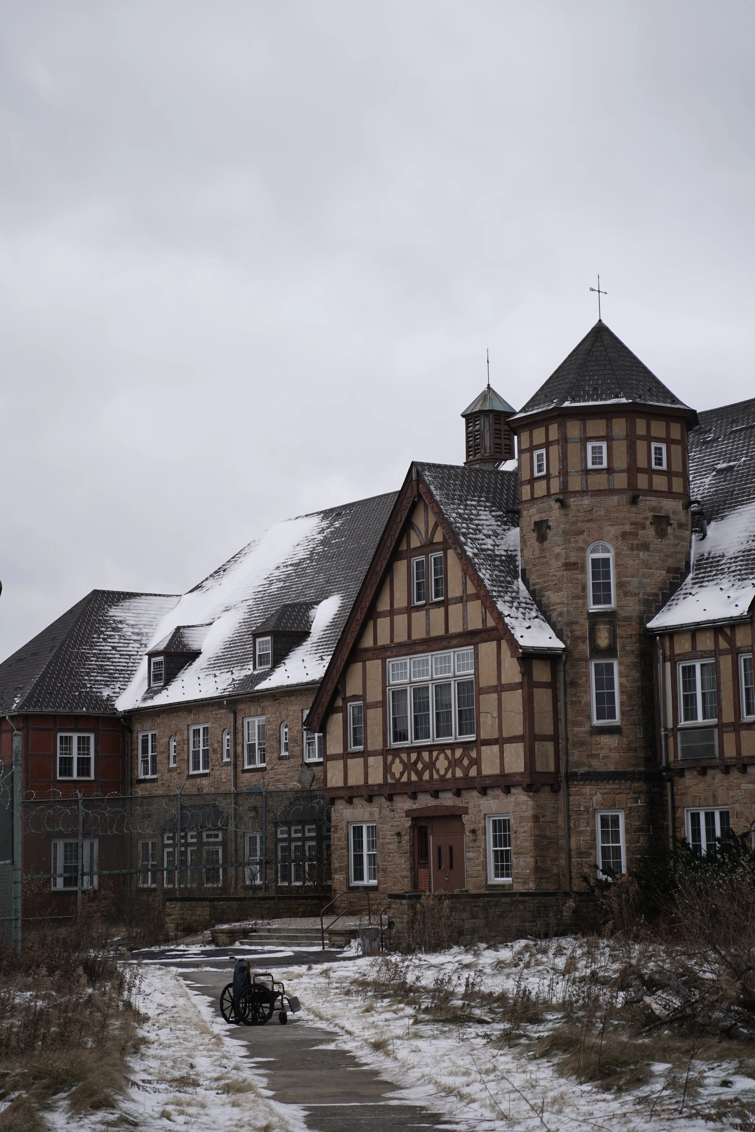 A snowy pathway leading to a historic stone and timber building with multiple rooflines and a tower, overcast sky in the background.