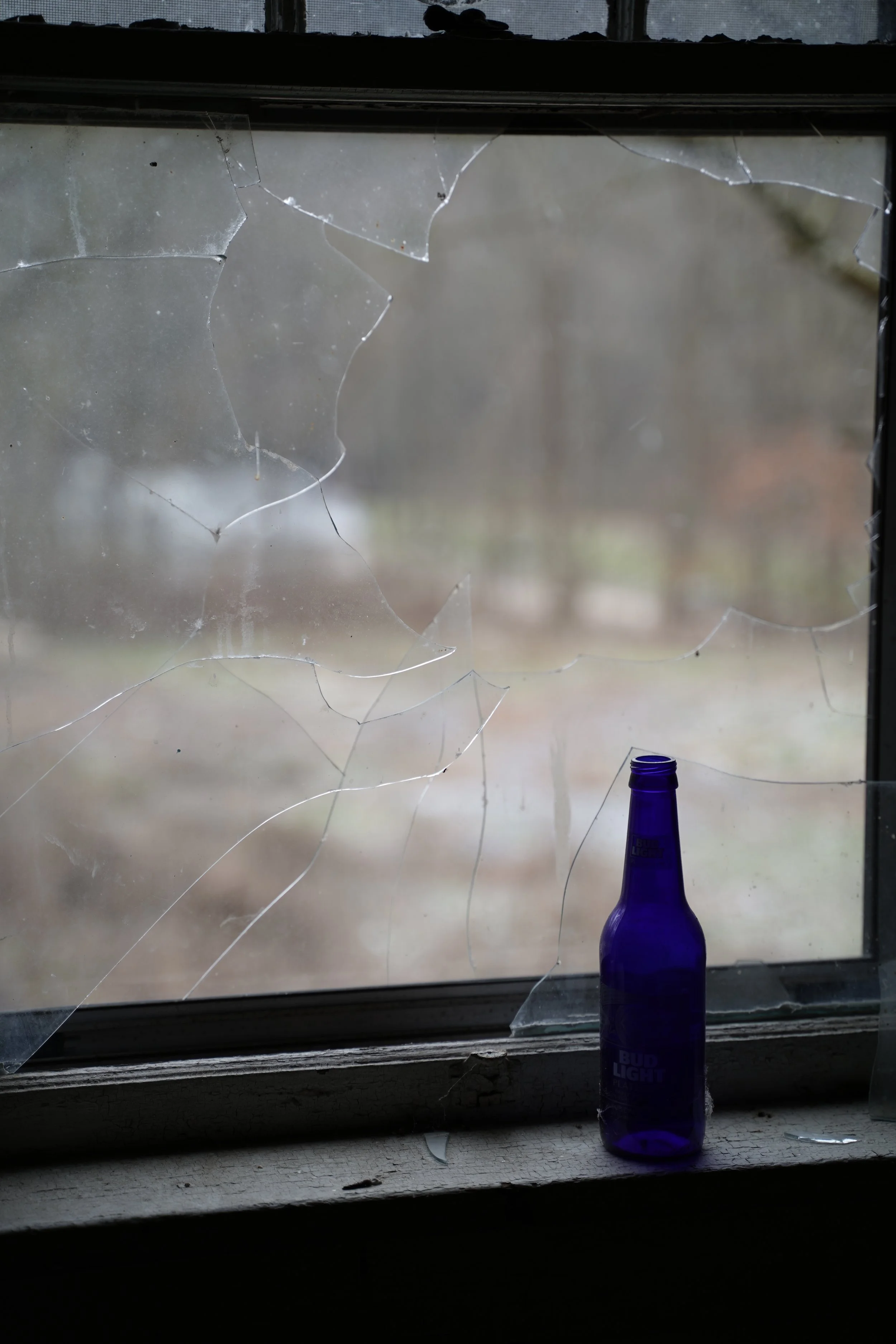 A broken window with shattered glass pieces, and a blue Bud Light beer bottle on the windowsill.