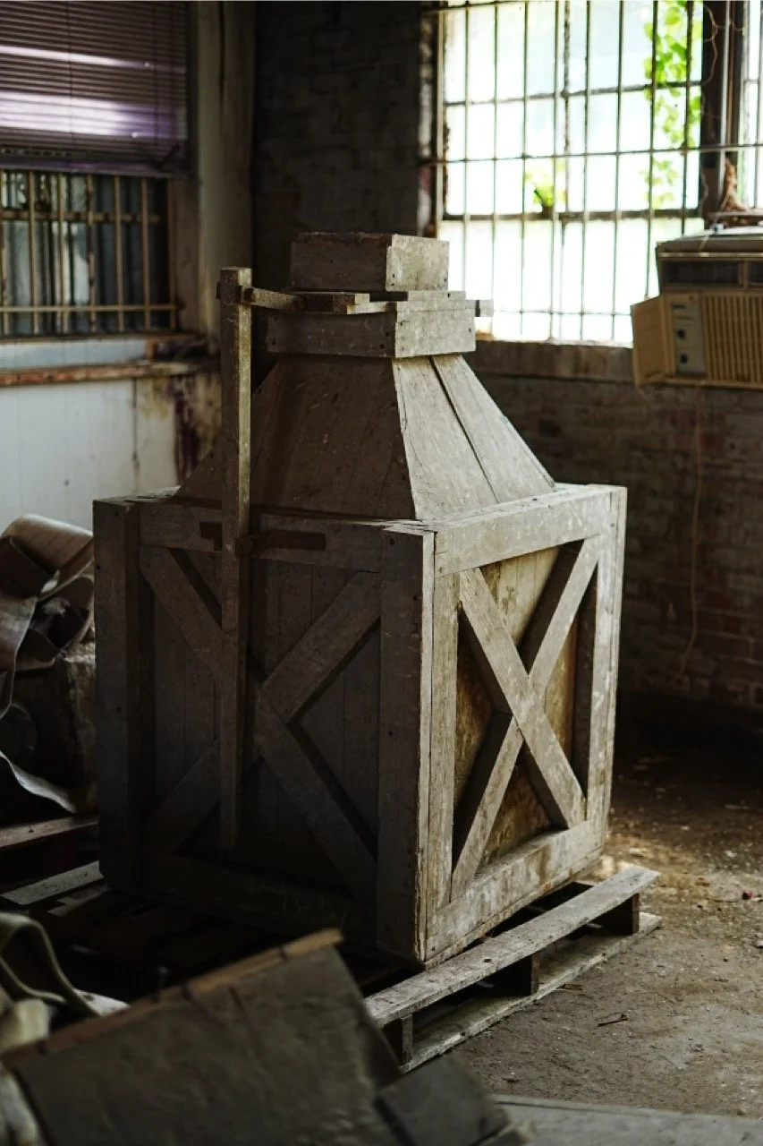 A large wooden crate with a conical top, sitting inside a rustic, possibly abandoned building with brick walls and old windows, illuminated by natural light.