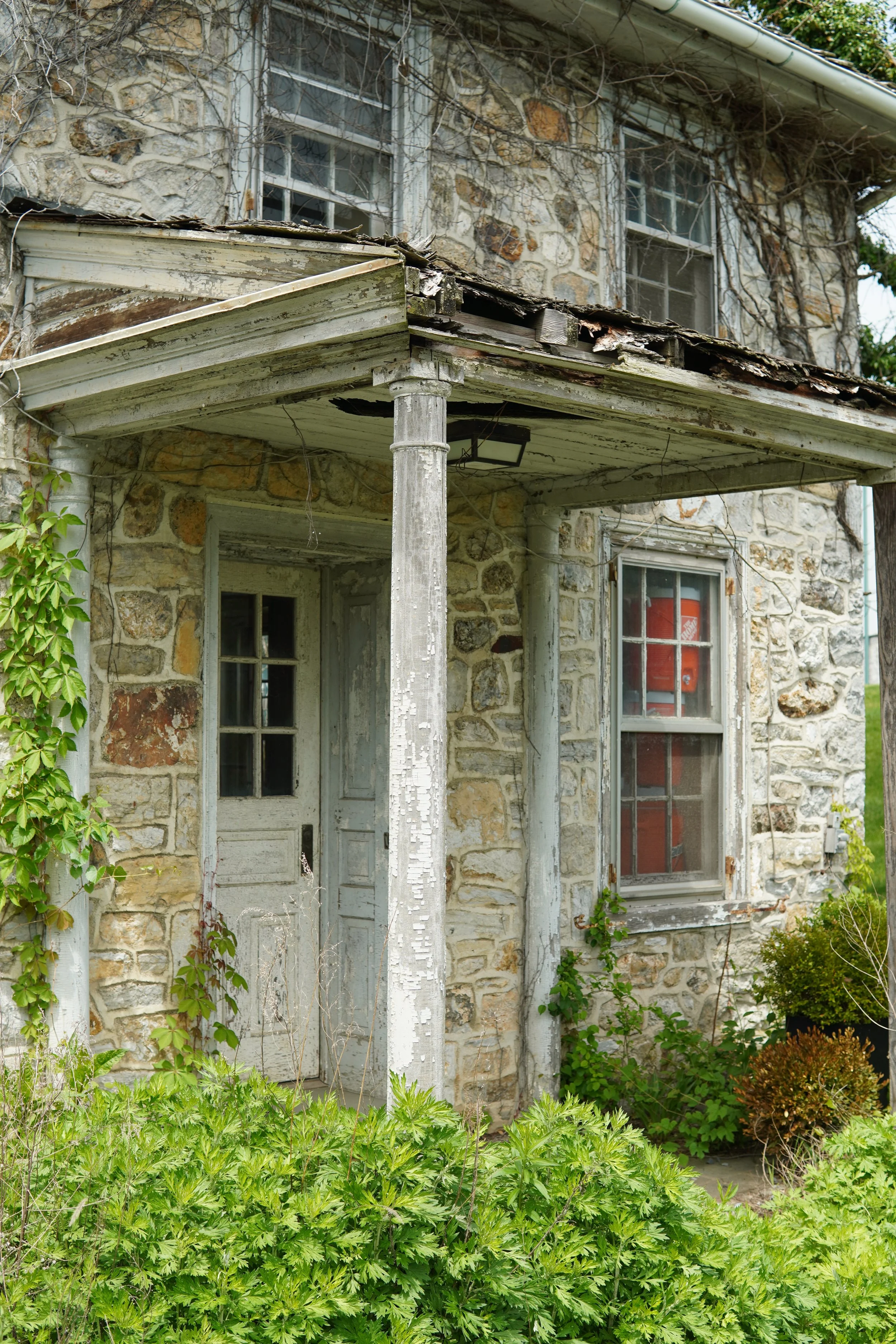 An old stone house with peeling white trim and a small porch, surrounded by overgrown plants and greenery.