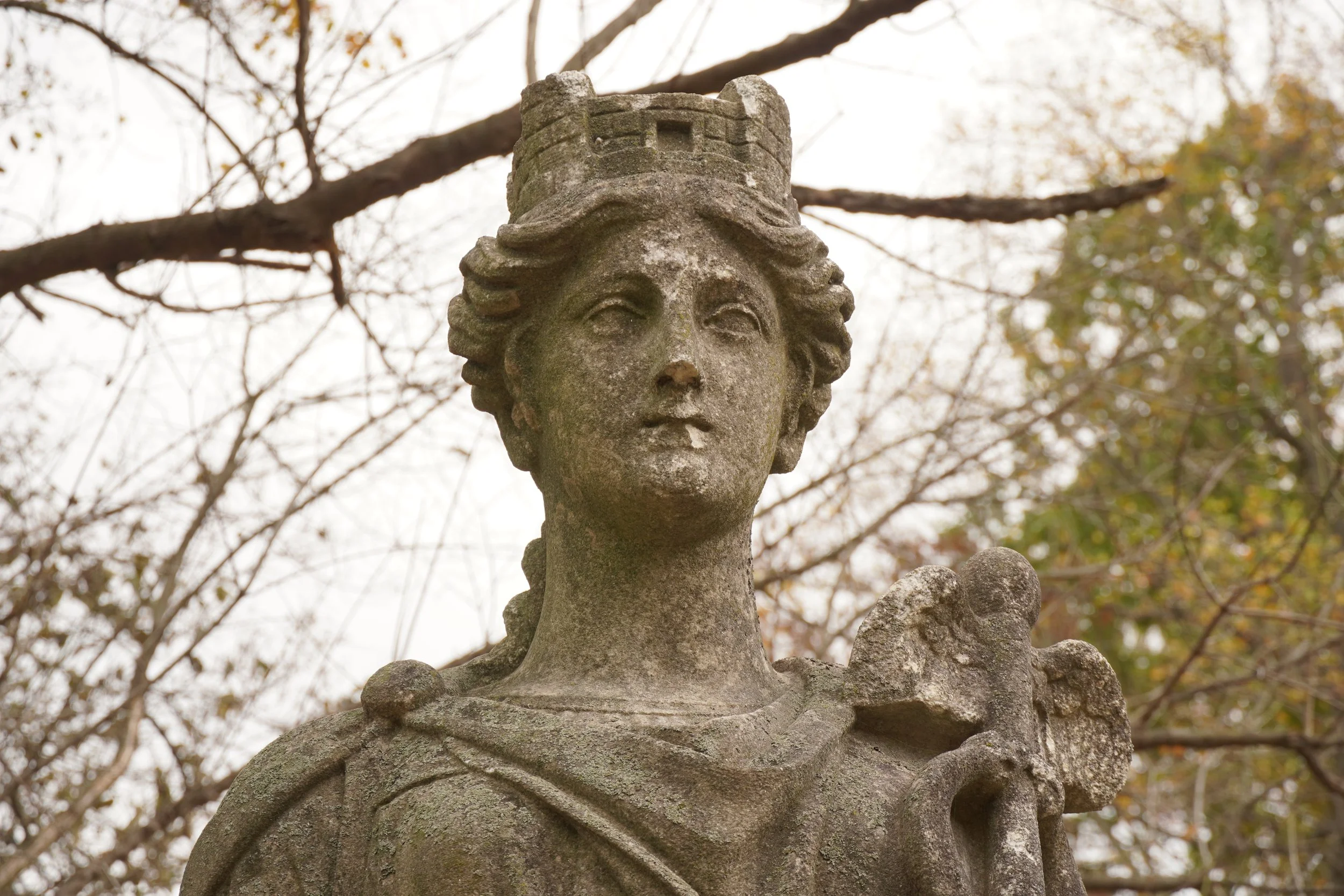 Close-up of a weathered stone statue of a woman with a small castle on her head, set outdoors with autumn trees in the background.