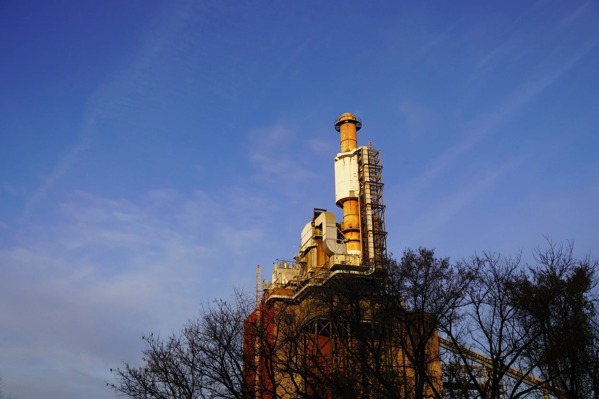 An industrial smokestack with metal structures and ladders, surrounded by leafless trees under a clear blue sky.