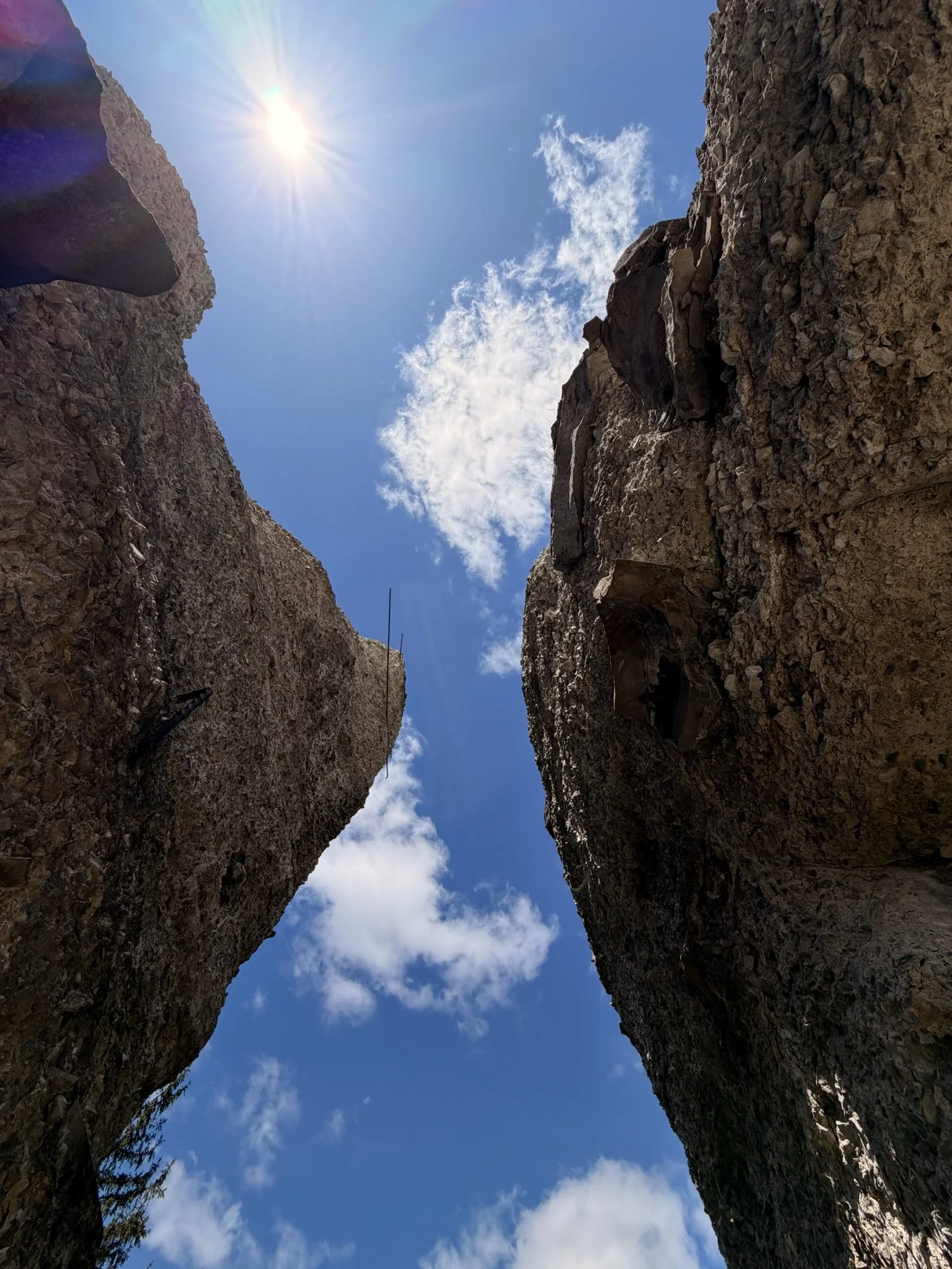 Looking up between two tall, rocky canyon walls to a bright sun and scattered white clouds in a blue sky.