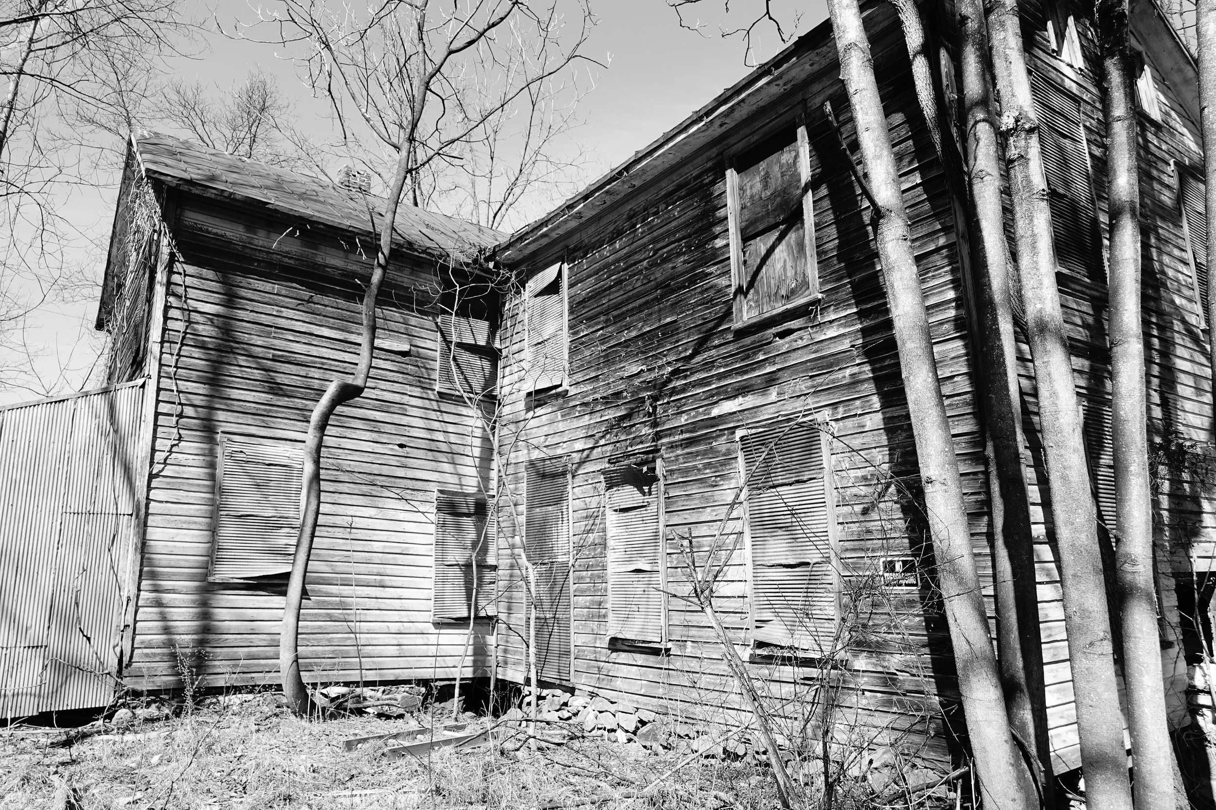 An old, weathered wooden house with broken windows and boarded-up openings, surrounded by leafless trees and overgrown weeds.
