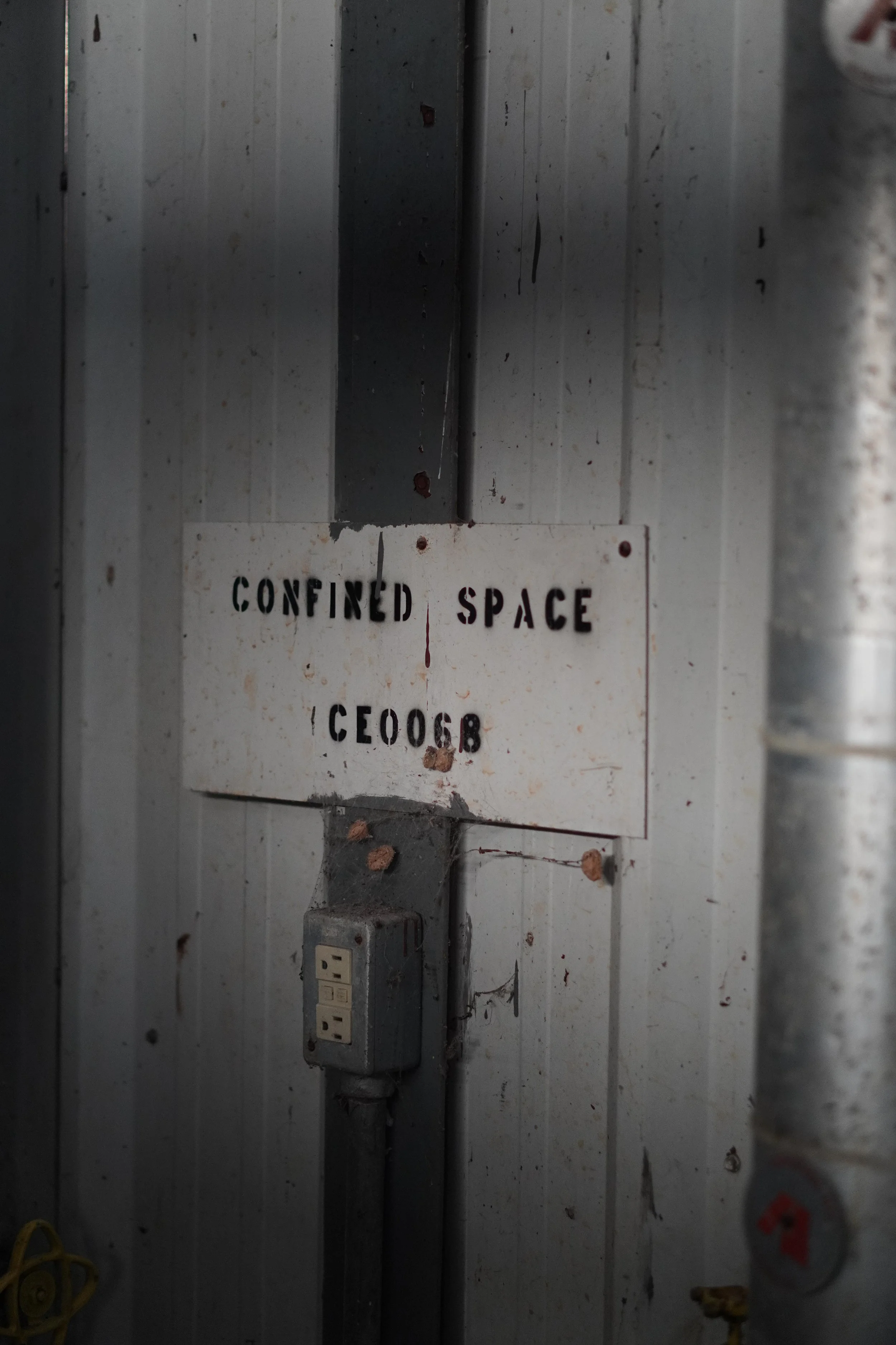 An old electrical panel with a warning sign that reads 'Contained Space' and a metal electrical box with outlets, mounted on a worn, dirty wall with visible grime and cobwebs.