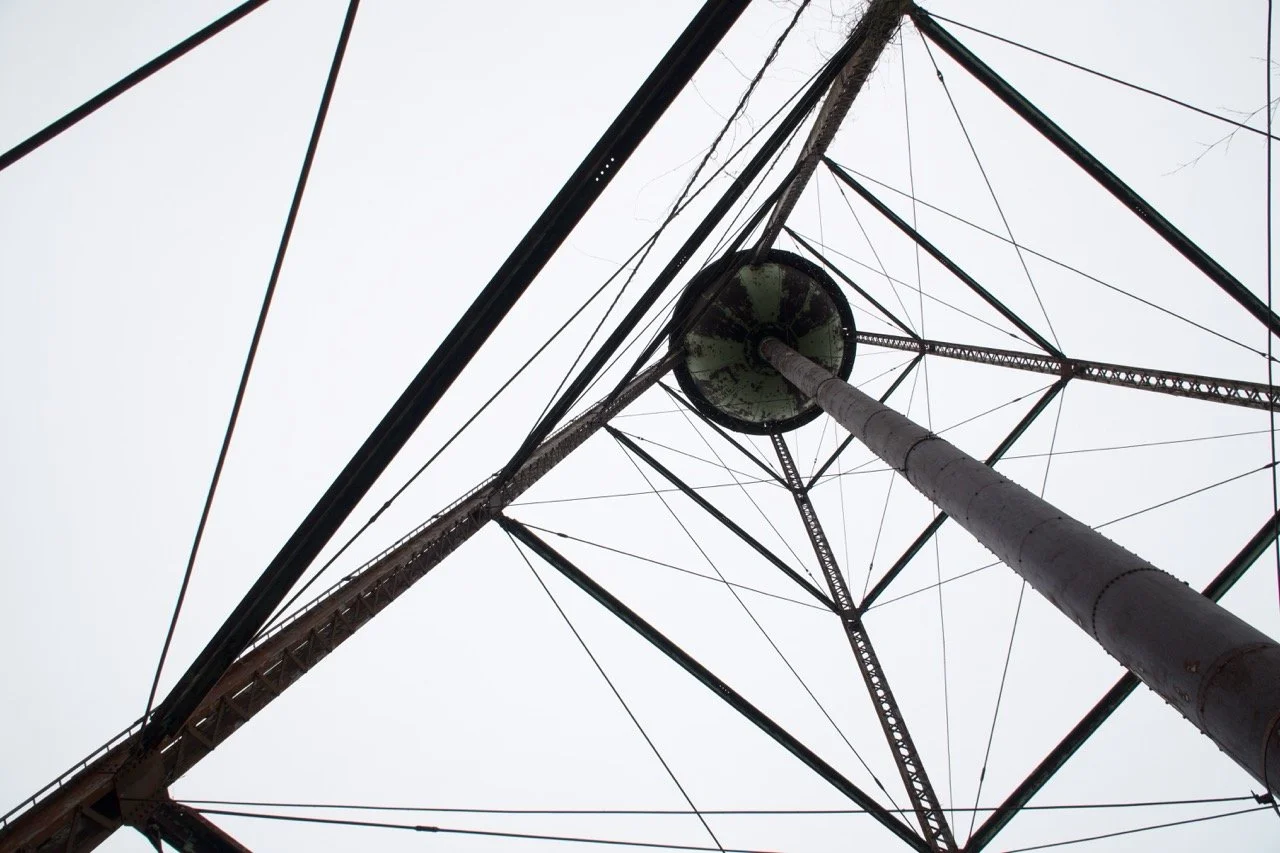 Looking up at the underbelly of a tall metal radio or communication tower with a large circular platform at the top, crisscrossed with numerous cables and supports against an overcast sky.