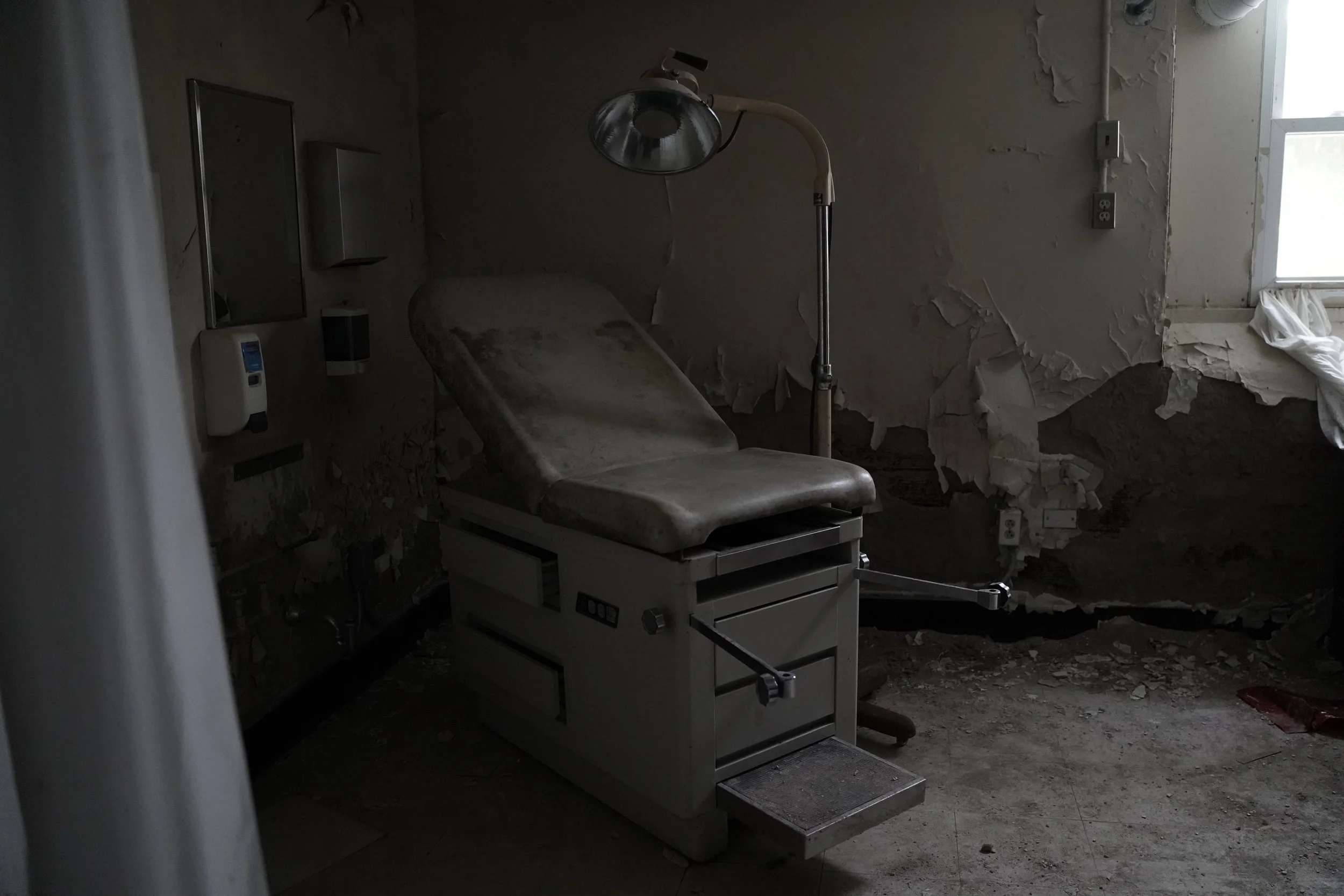 An abandoned hospital or medical room with a peeling wall, a rusty examination table, and broken windows letting in natural light.