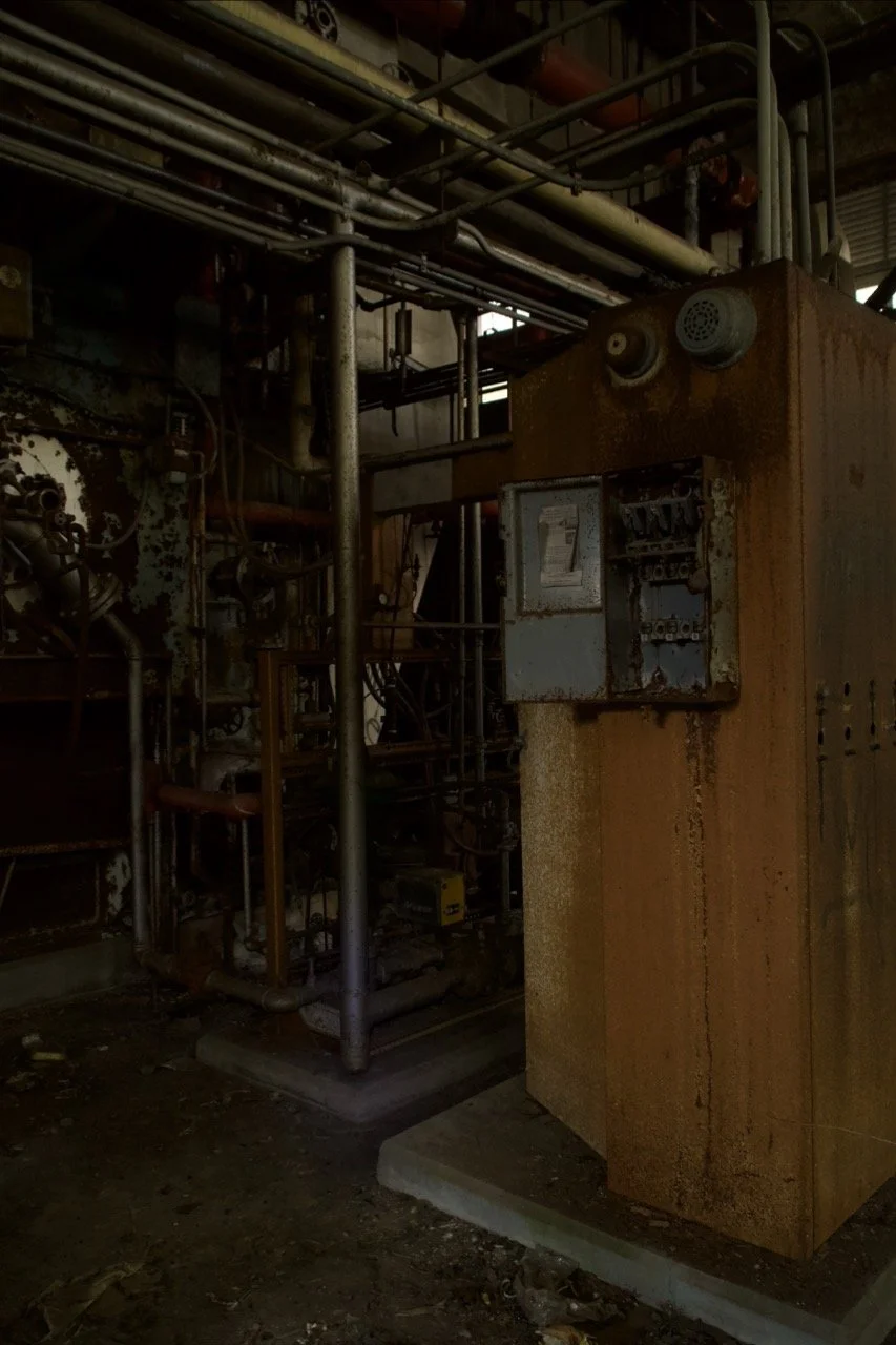 An old, rusty industrial electrical control panel with pipes and wires surrounding it in a dimly lit environment.