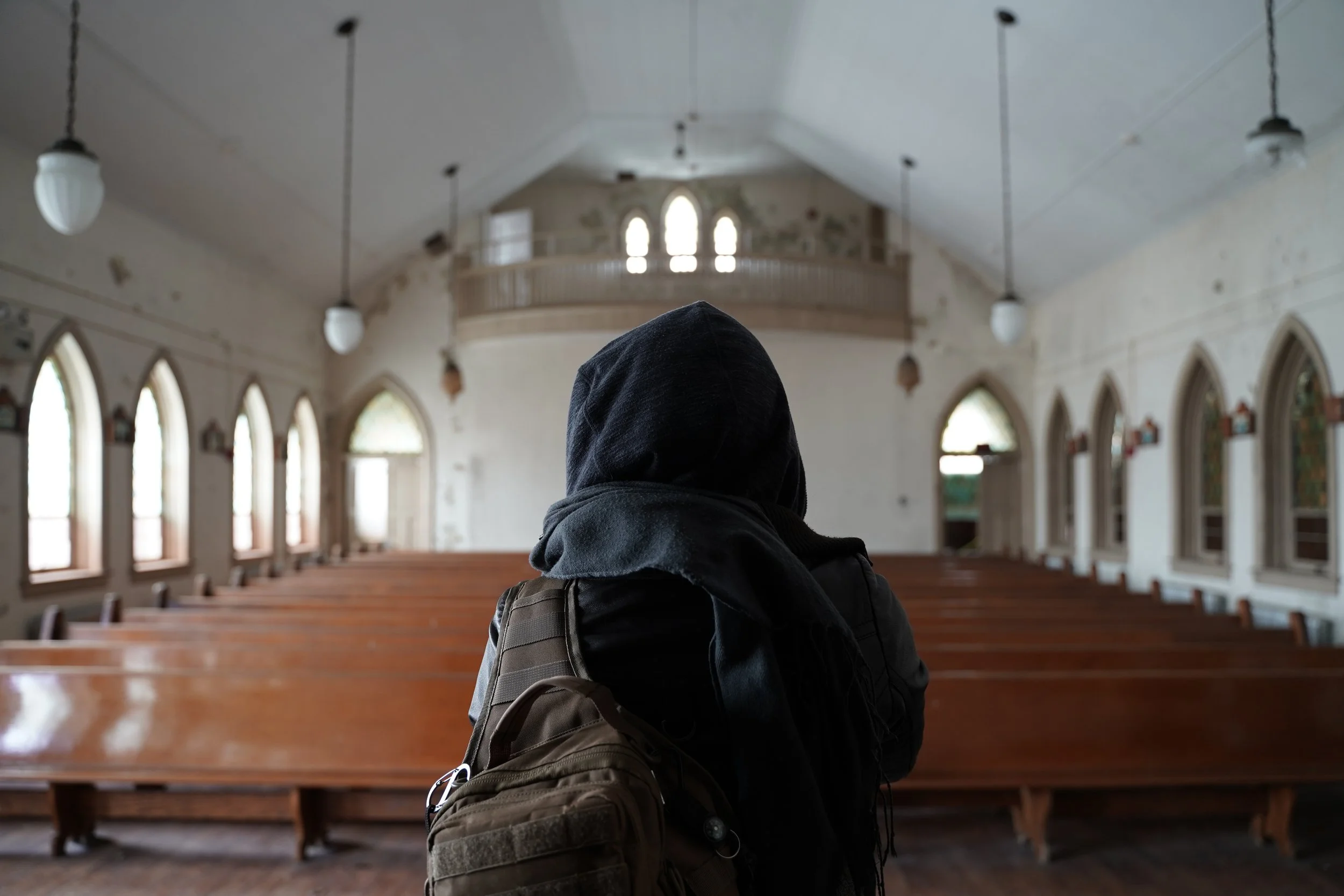 A person wearing a hoodie and carrying a backpack inside an empty church with wooden pews and stained glass windows.