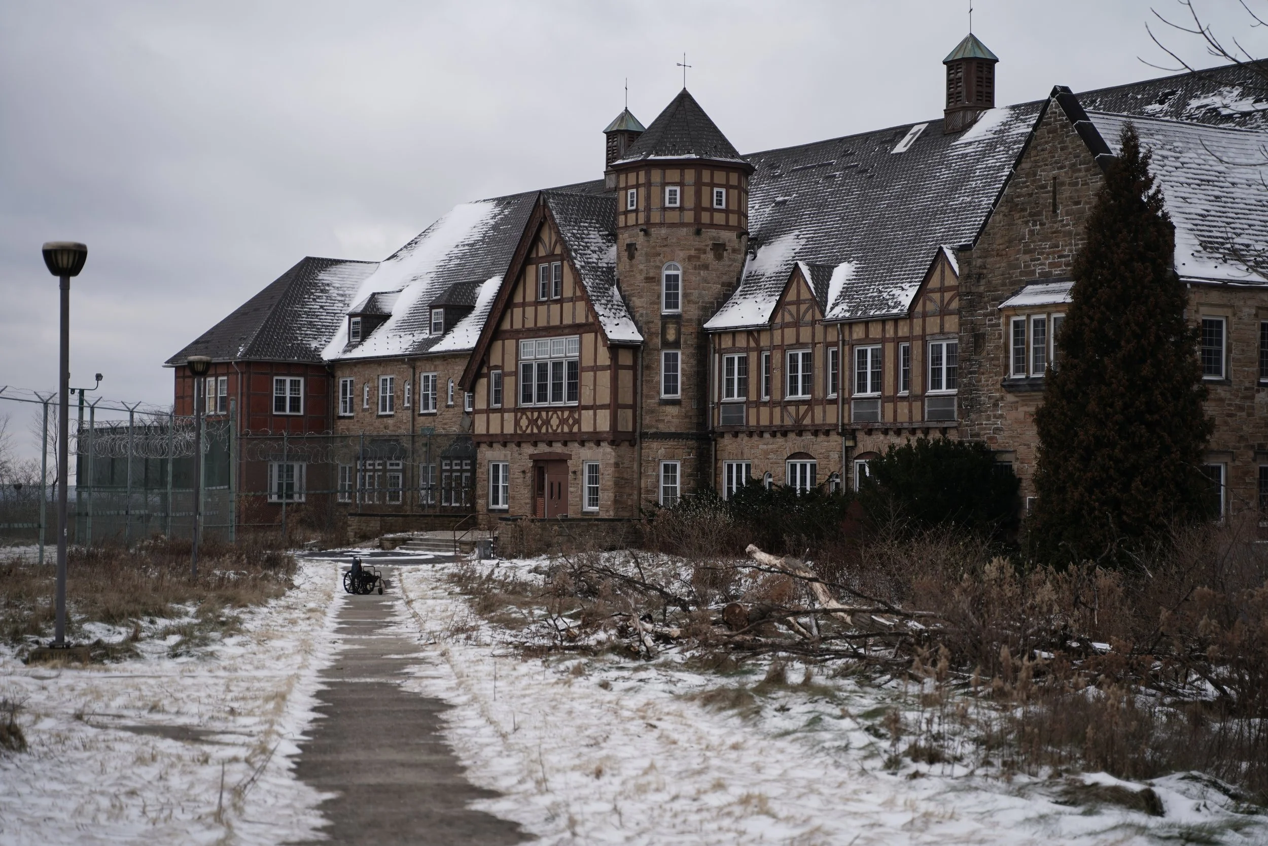A large, old stone and timber building with multiple steep roofs partially covered in snow, surrounded by a chain-link fence, with a cloudy sky above and a wheelchair on a sidewalk in the foreground.