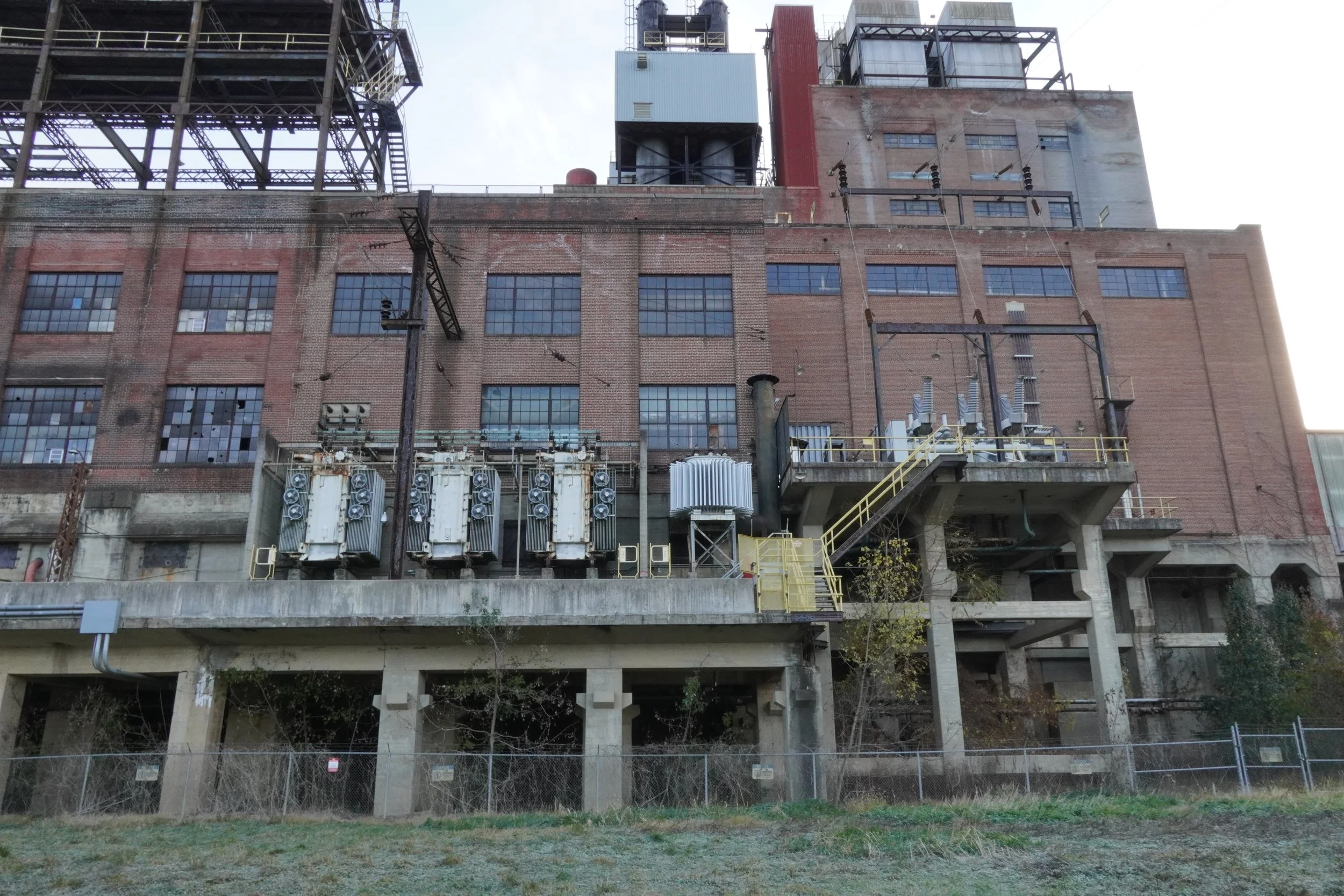 The back side of an industrial building with large windows, electrical equipment, and structural supports, viewed from street level with a chain-link fence in the foreground.