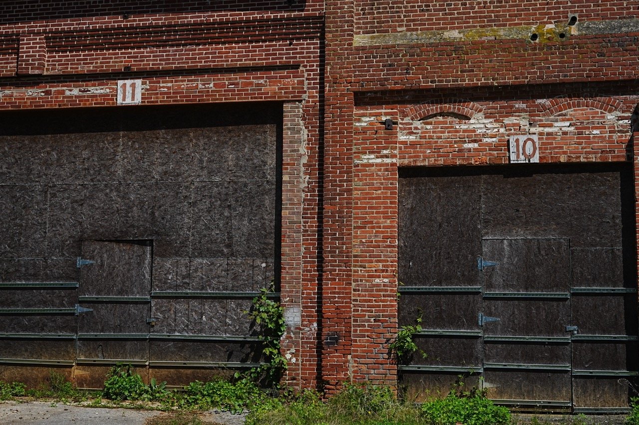 Two boarded-up brick building doors, one with the number 11 and the other with 10, separated by a brick column, with some overgrown plants at the bottom.