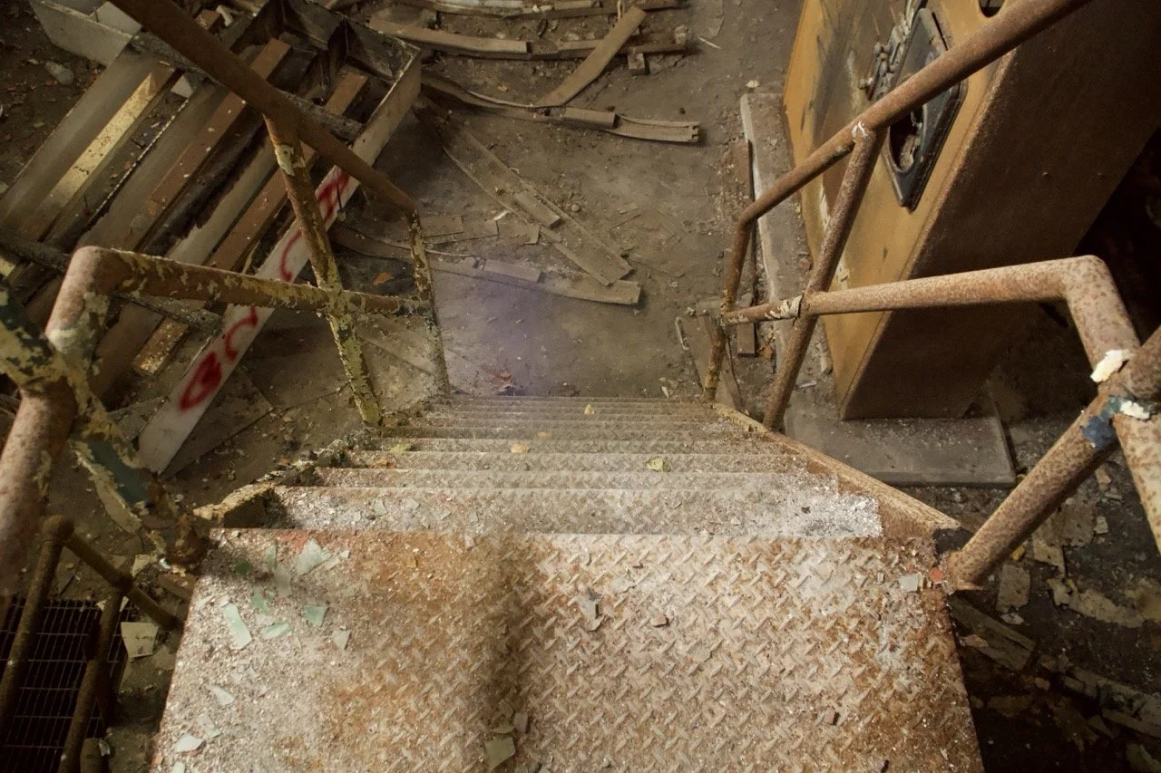 View of a rusty metal staircase descending into an abandoned, dusty, and debris-filled space, with chipped and peeling paint on the railing.