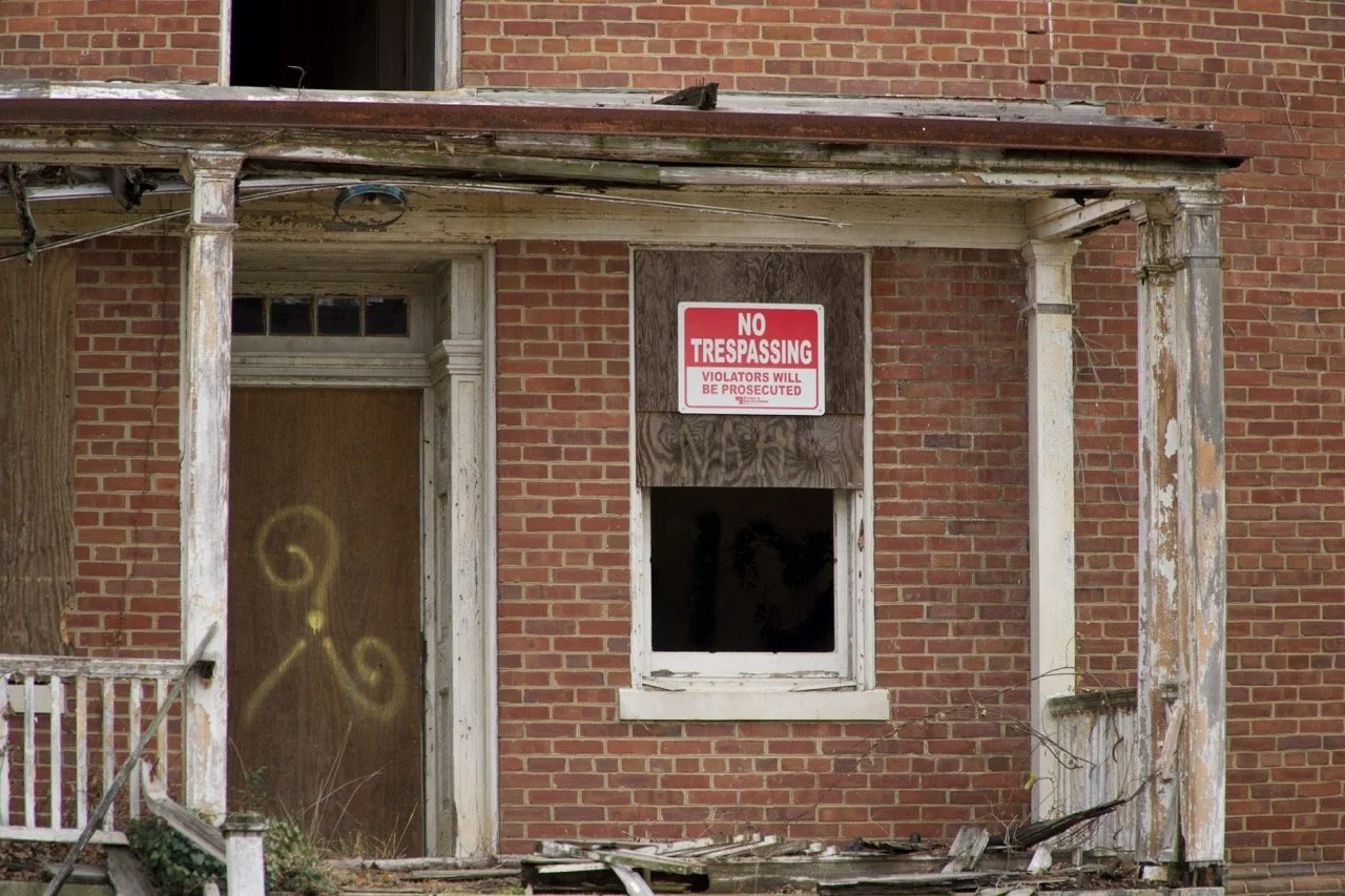 Abandoned brick house with a torn porch, boarded-up window covered with plywood, and a 'No Trespassing' sign on the wall.