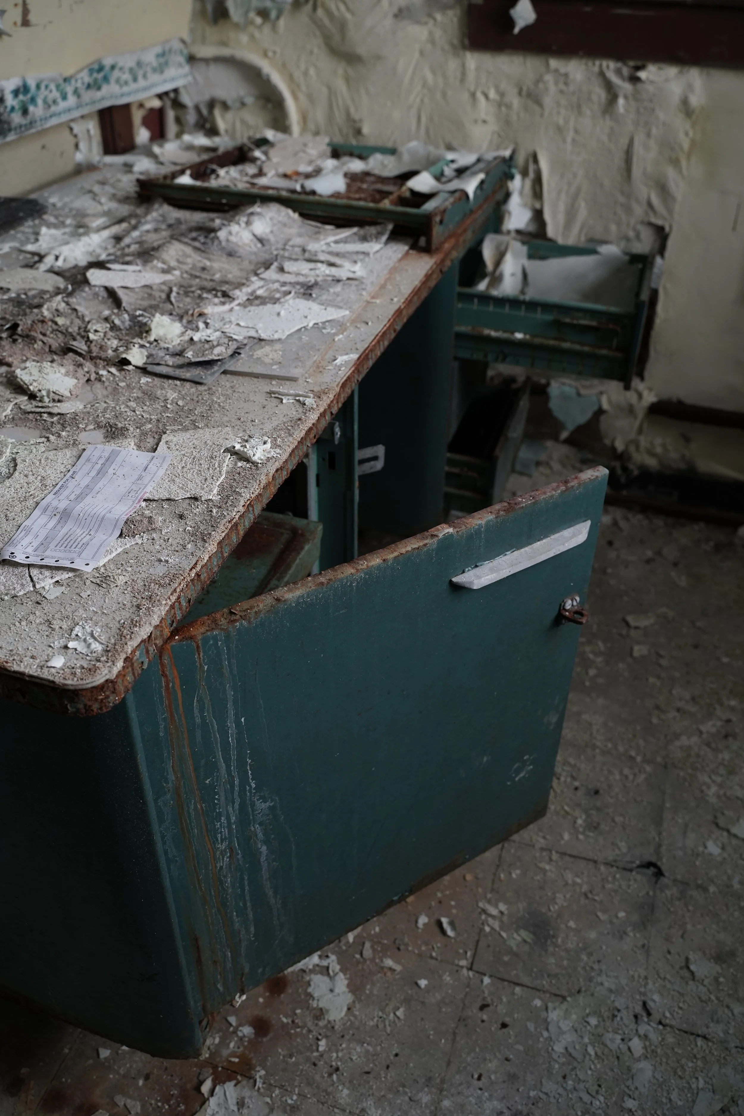 A devastated room with a rusty trash bin filled with debris and torn papers, dirt, and rubble scattered across the floor and a broken wall with damaged plaster.
