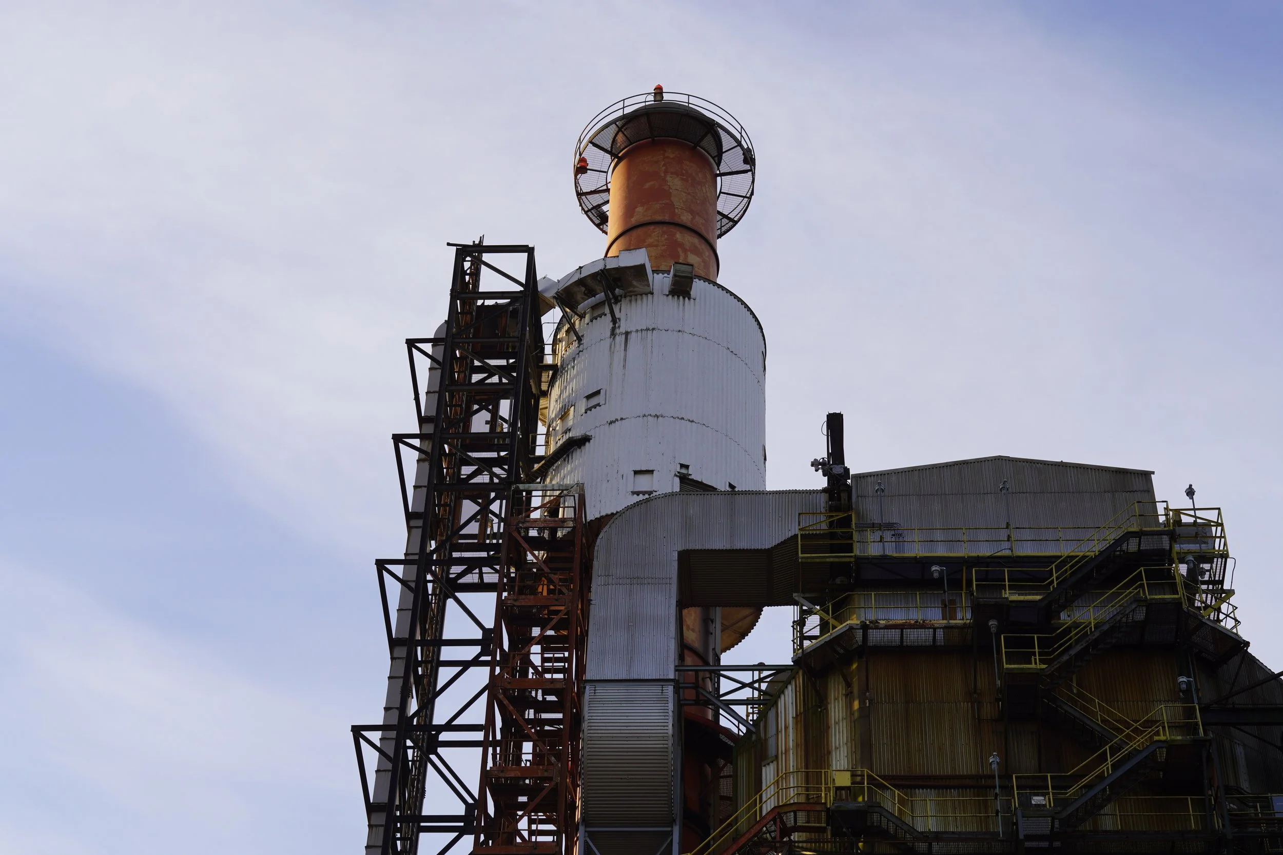 An industrial factory chimney with metal stairs and yellow safety railings against a clear sky.