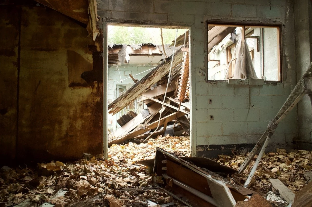 An abandoned room with debris and fallen leaves on the floor, a broken wall, and an open door revealing a collapsed roof outside.