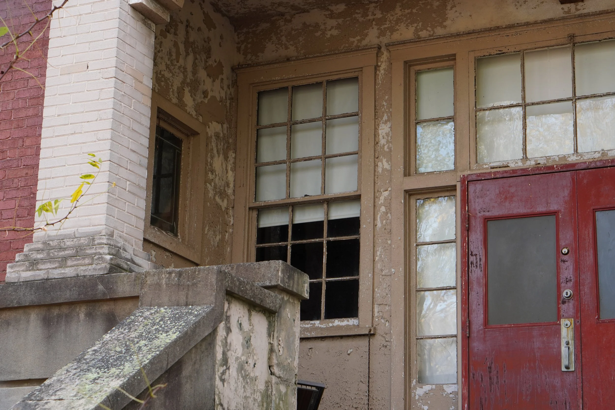 An old building with peeling paint, cracked walls, and a weathered red door. The building has large multi-pane windows with dirt and grime on the glass, showing signs of neglect and decay.
