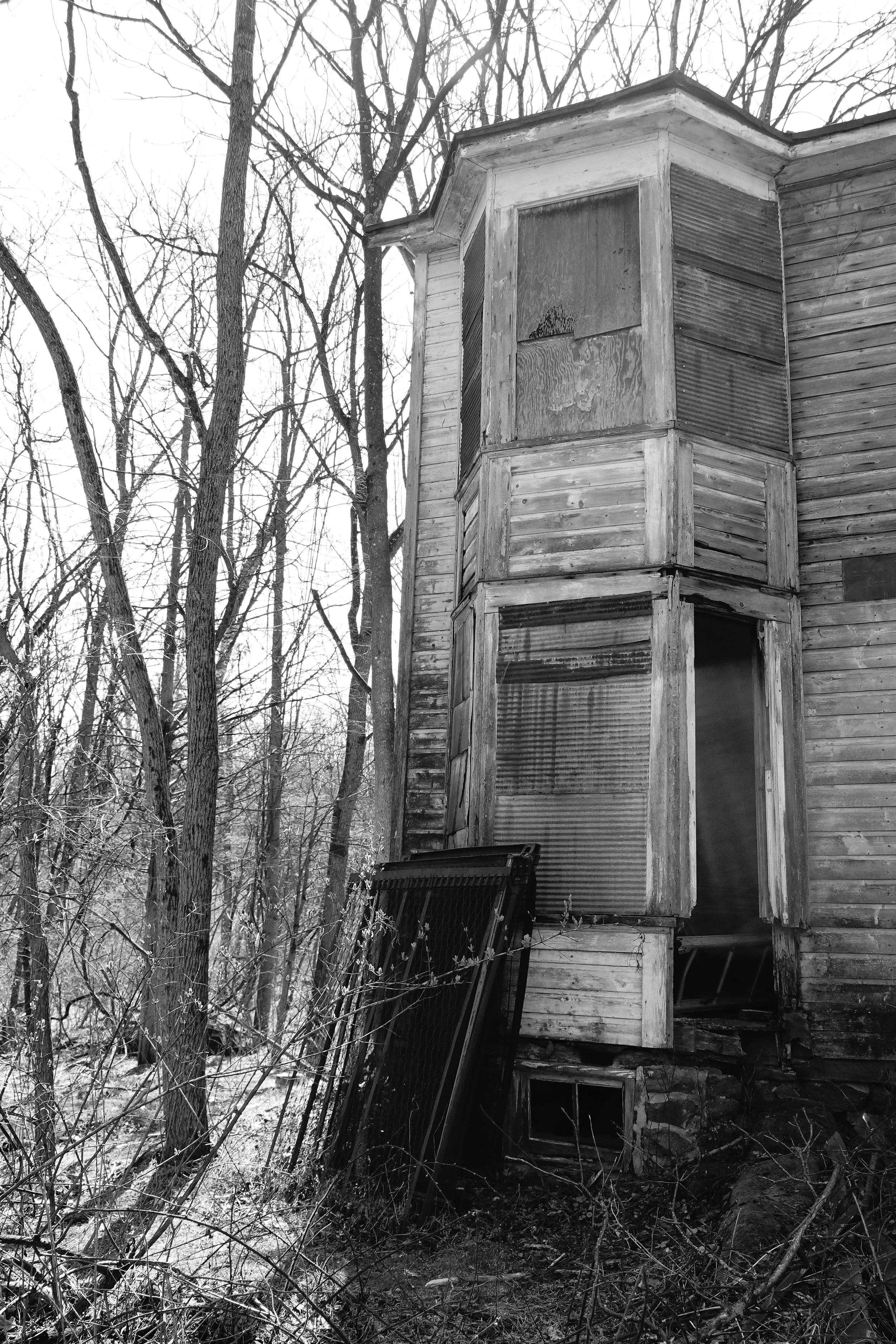 An old, abandoned wooden house in a wooded area with leafless trees. The house has boarded-up windows and peeling paint, indicating it has been neglected for some time.
